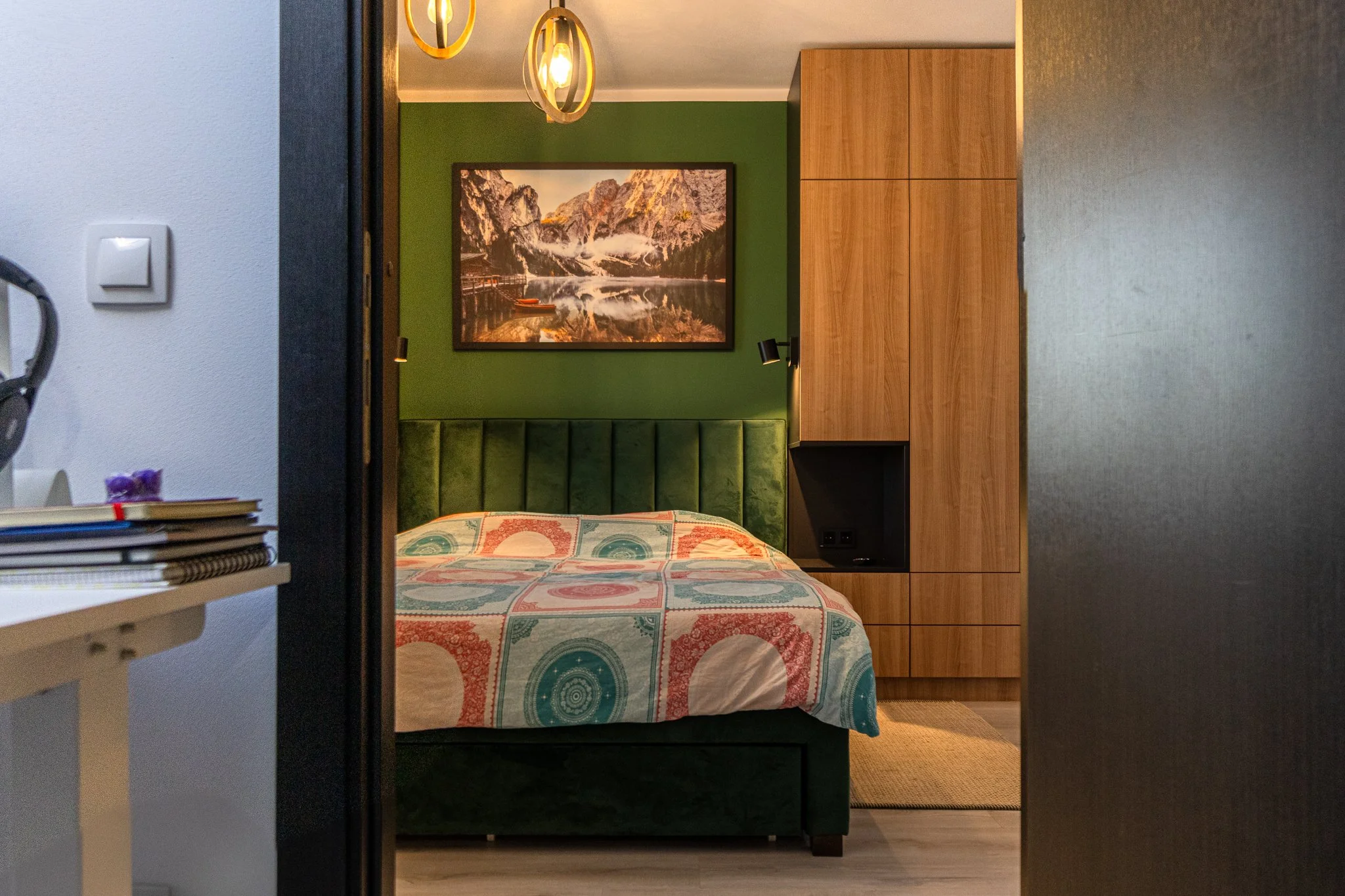 View into a bedroom with a green upholstered headboard, a colorful bedspread, a landscape painting of mountains and a lake on a green wall, and a tall wooden wardrobe. Part of a white table with books and a purple object is visible on the left.