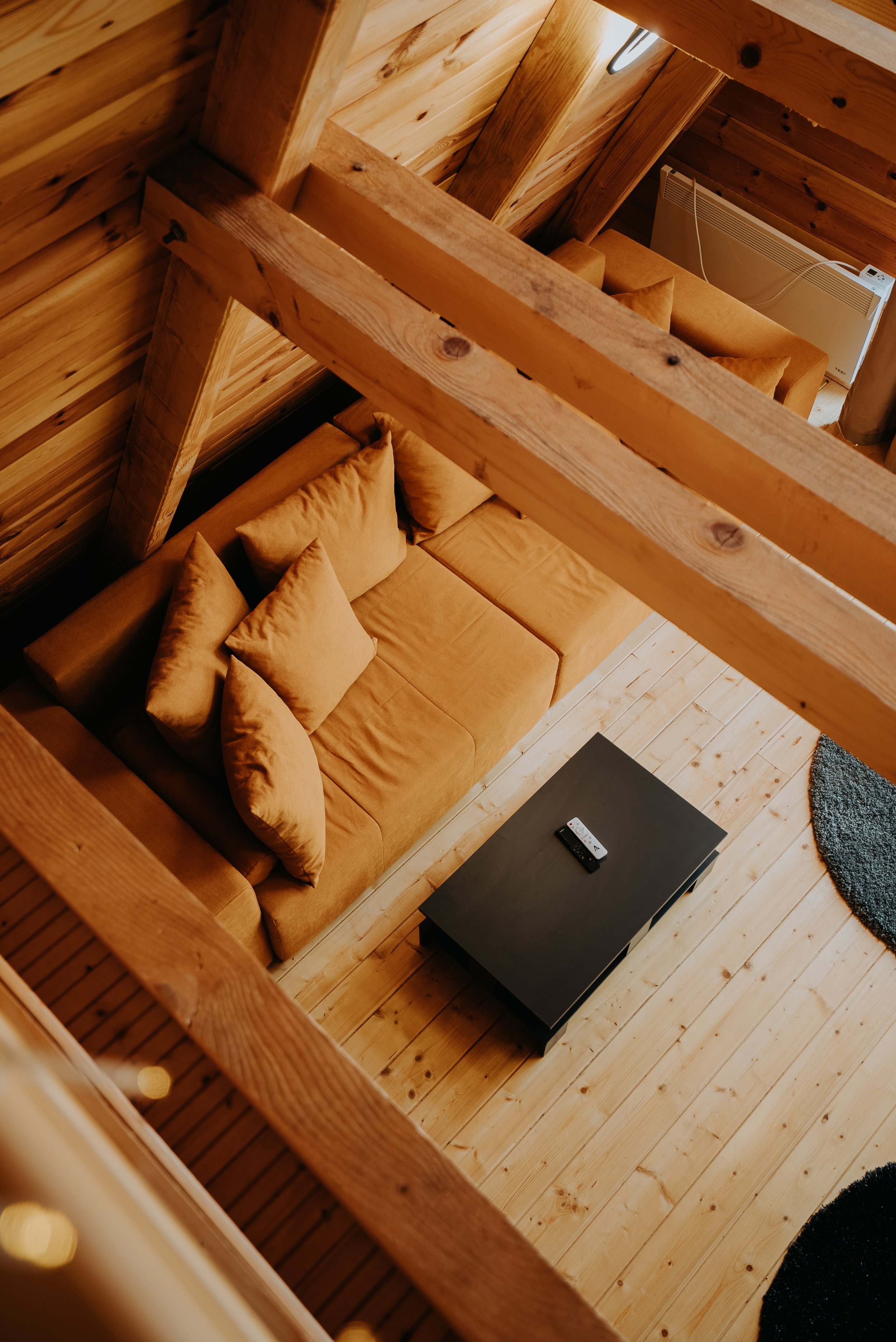 View of a cozy living room from above, with a tan couch, black coffee table with a remote, and a mounted heater, all inside a wooden cabin with visible beams and flooring.