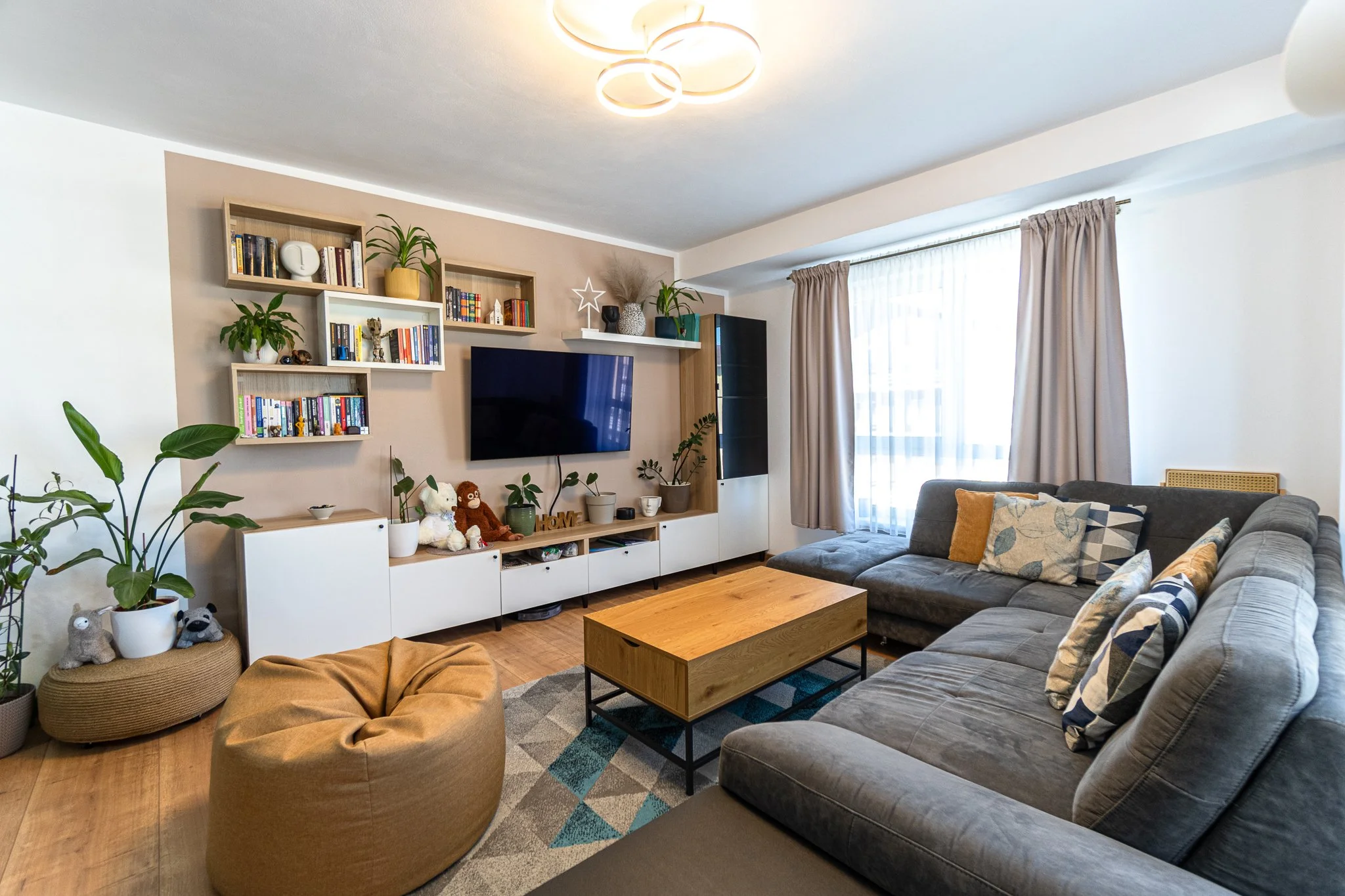 Living room with a large gray sectional sofa, beige curtains, a wooden coffee table, and a media console with a mounted flat-screen TV. The wall behind the TV has shelves with books, plants, and decorative items. There are potted plants on the floor 