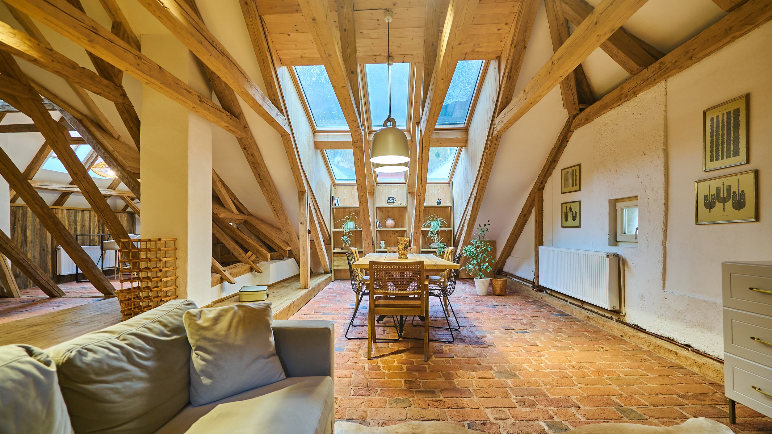 An attic dining area with exposed wooden beams, large skylight windows, a brick floor, a dining table with six chairs, and a built-in bookshelf at the far end.