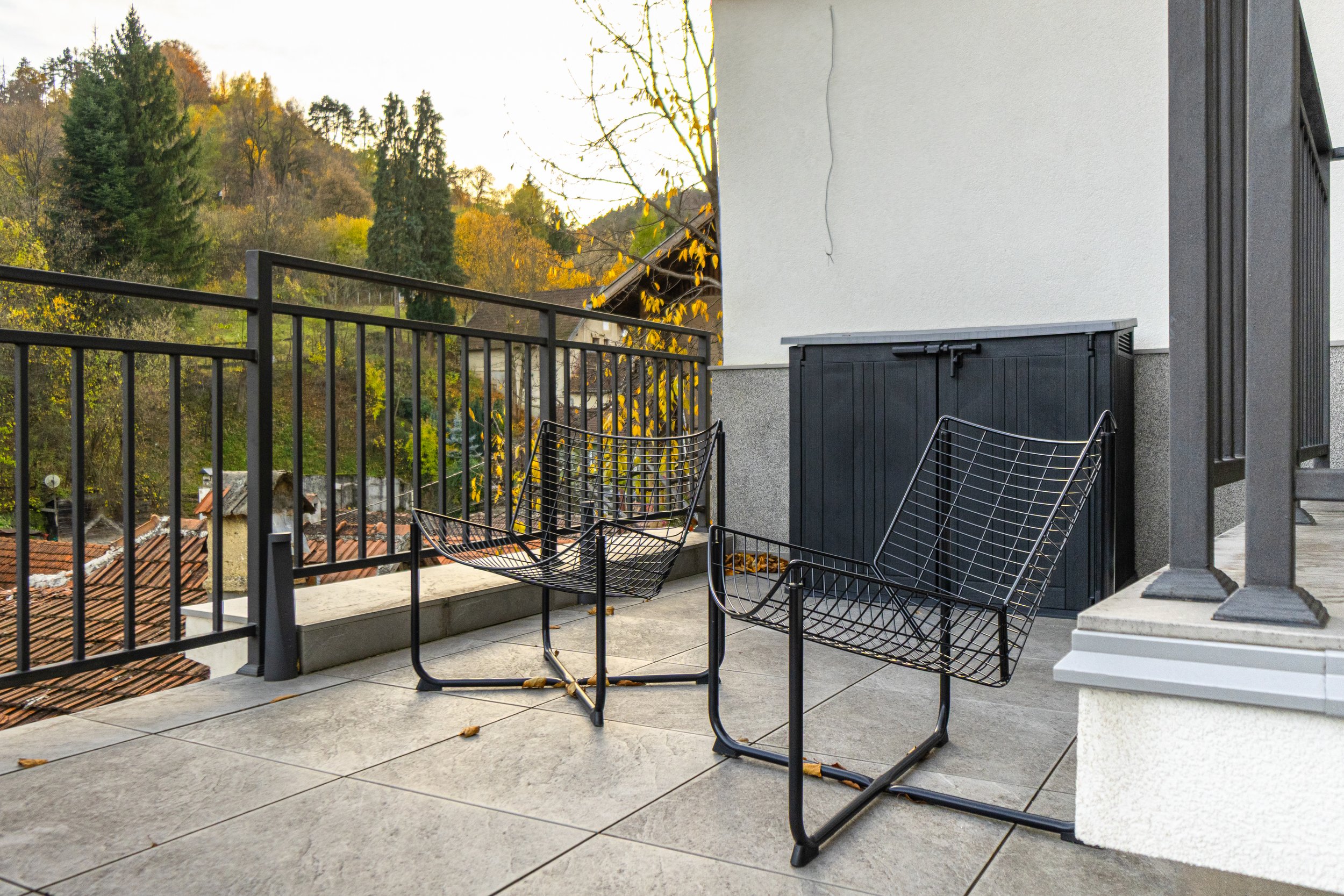 View of a balcony with two black wire chairs and a black storage box, with a landscape of trees with fall foliage in the background.