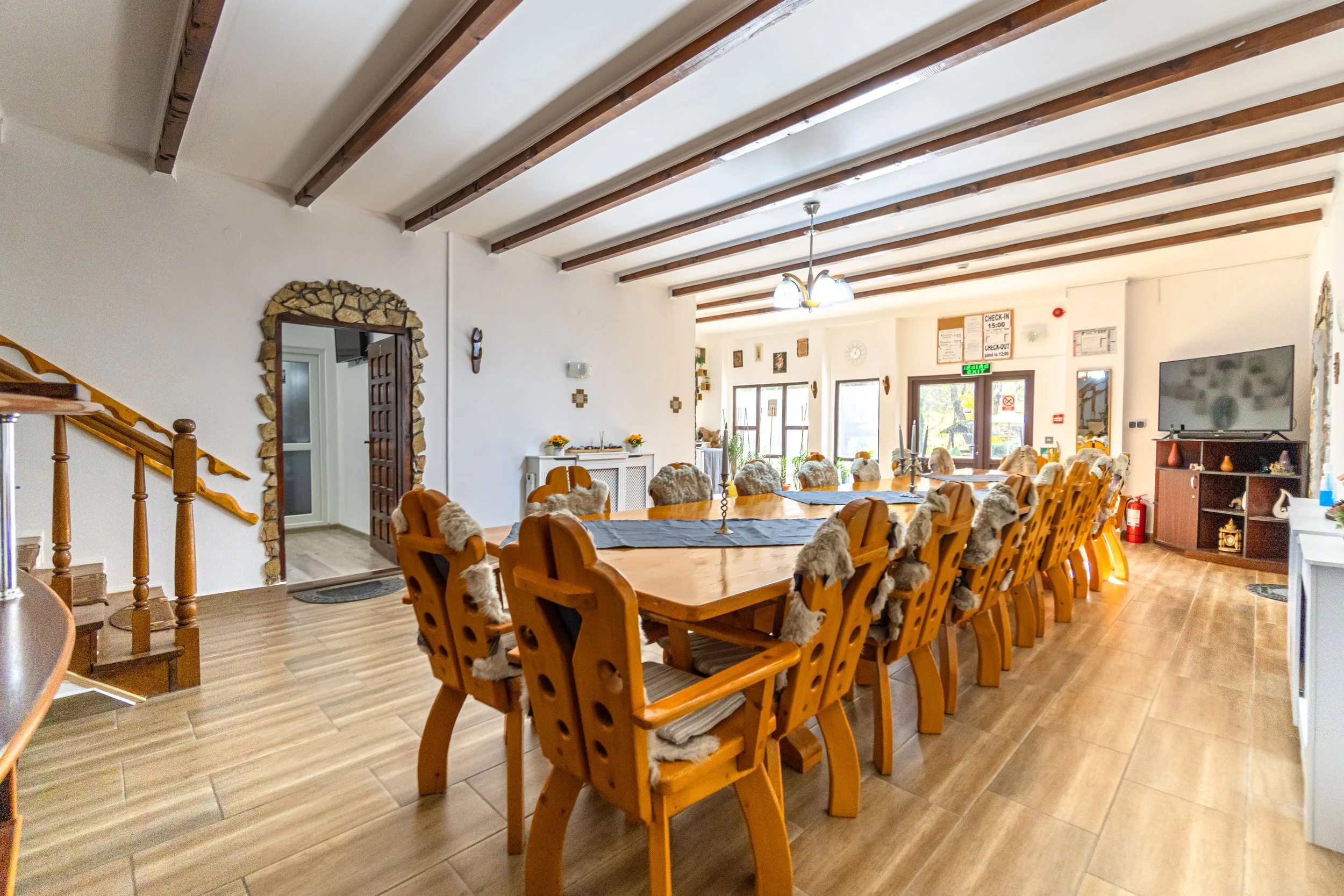 Large dining room with a long wooden table surrounded by chairs with fur cushions, a TV on a stand, and large windows letting in natural light.