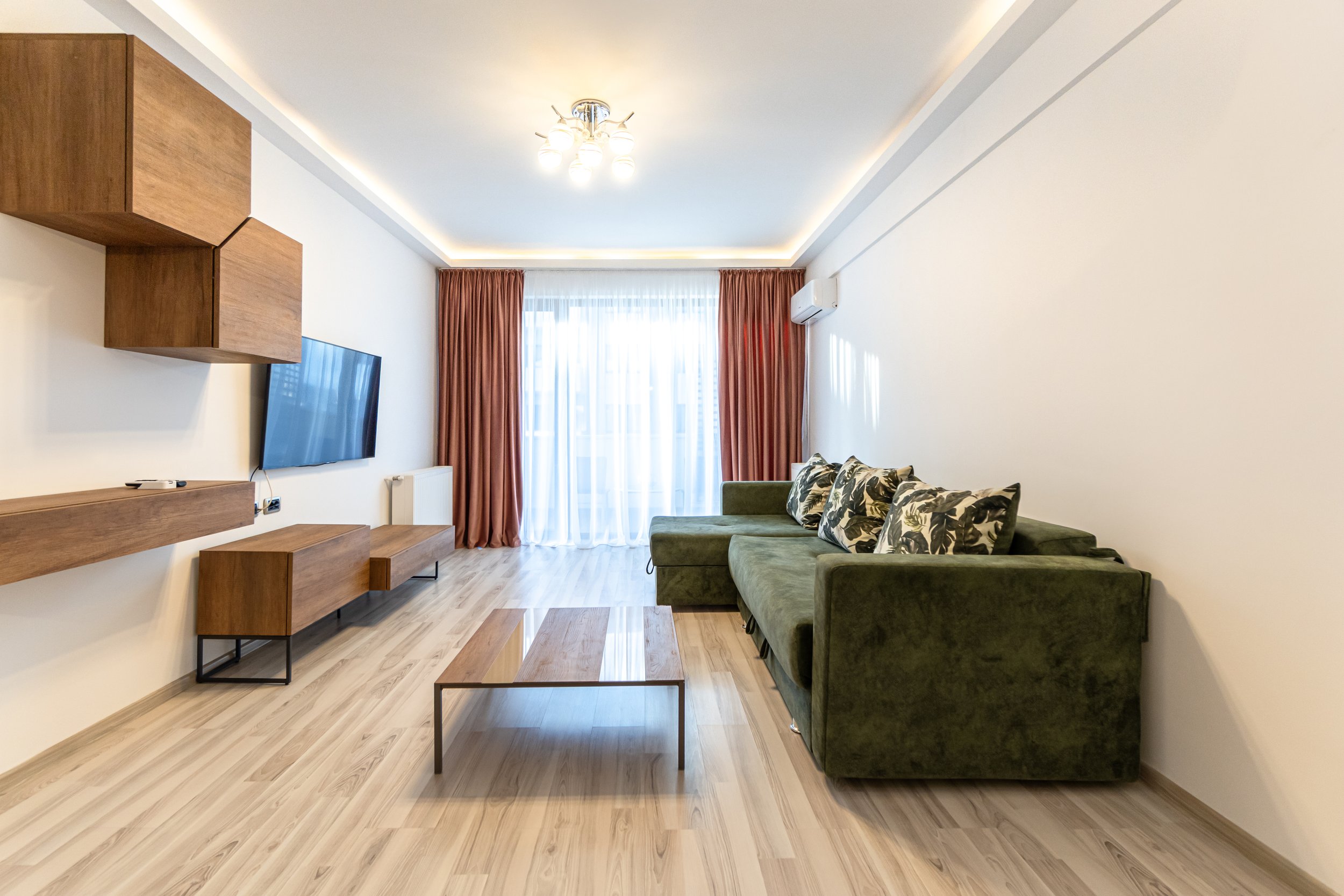 Modern living room with a green sectional sofa, wooden coffee table, wall-mounted TV, floating wooden shelves, curtains, and wood flooring.