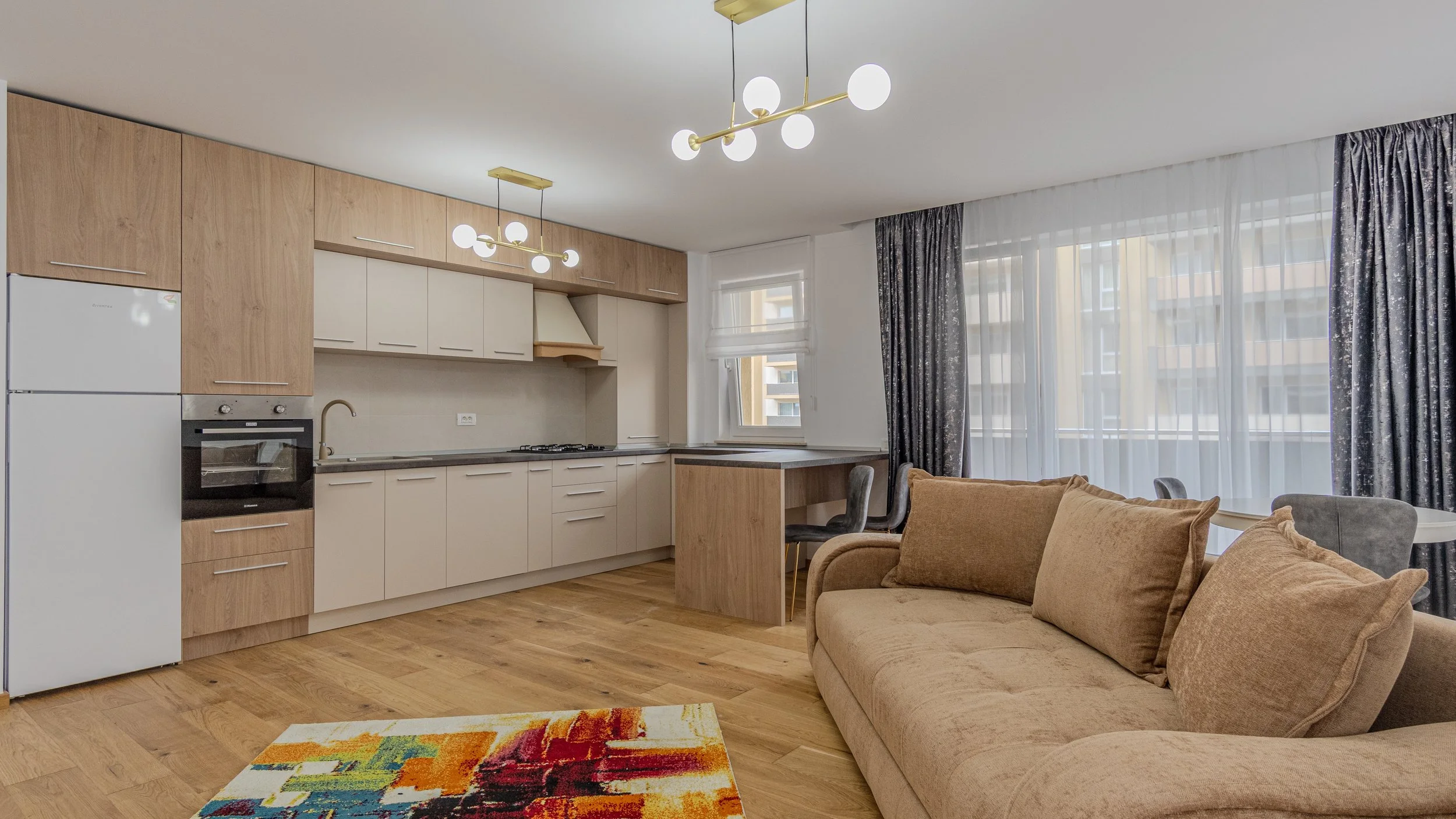 Living room and kitchen area with beige couch, colorful abstract rug, wooden flooring, large windows with sheer and dark curtains, modern lighting fixtures, and a compact kitchen with beige and wood cabinets, a built-in oven, sink, and countertop.