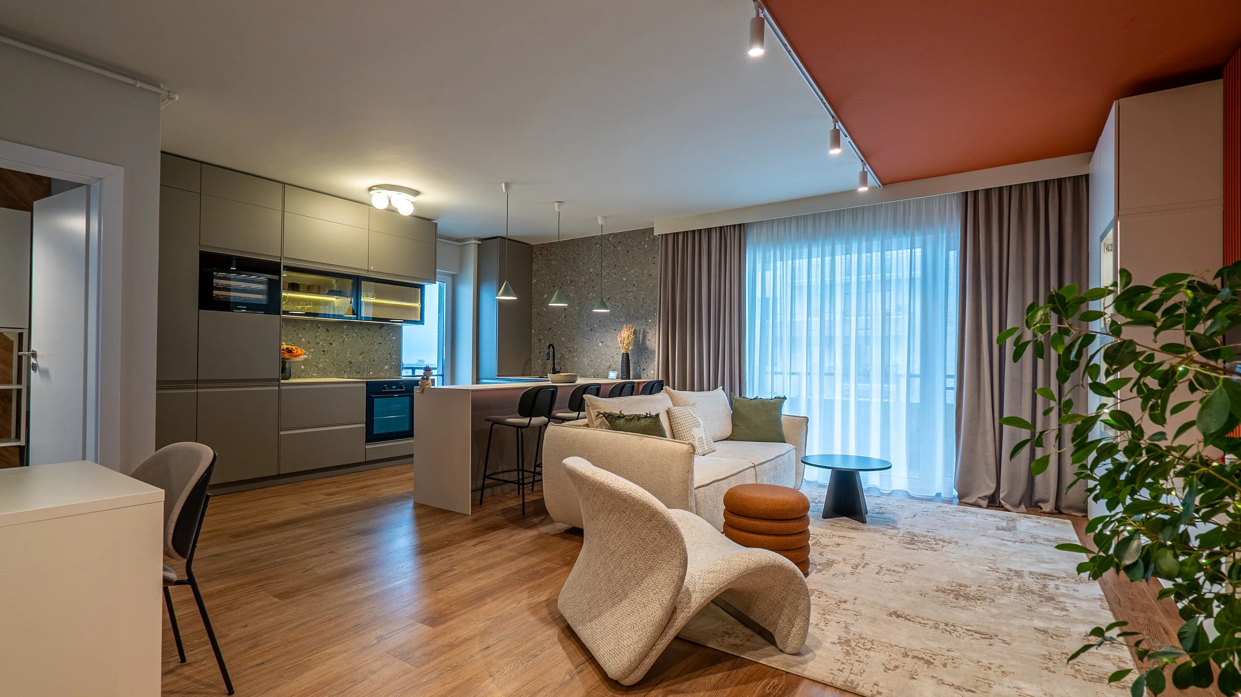 Modern open-concept living room and kitchen with beige sofa, textured armchair, barstools at kitchen island, large window with curtains, wooden flooring, indoor plant, and minimalist decor.