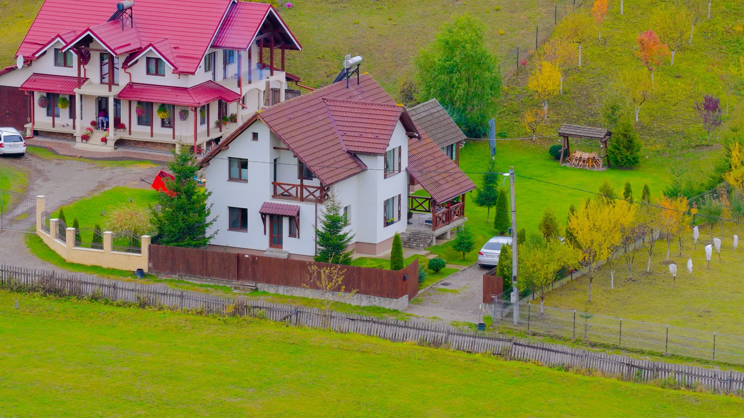 Two houses with red tiled roofs and white walls surrounded by green lawns, trees, and a wooden fence; one house has a balcony, and there are parked cars and outdoor furniture.