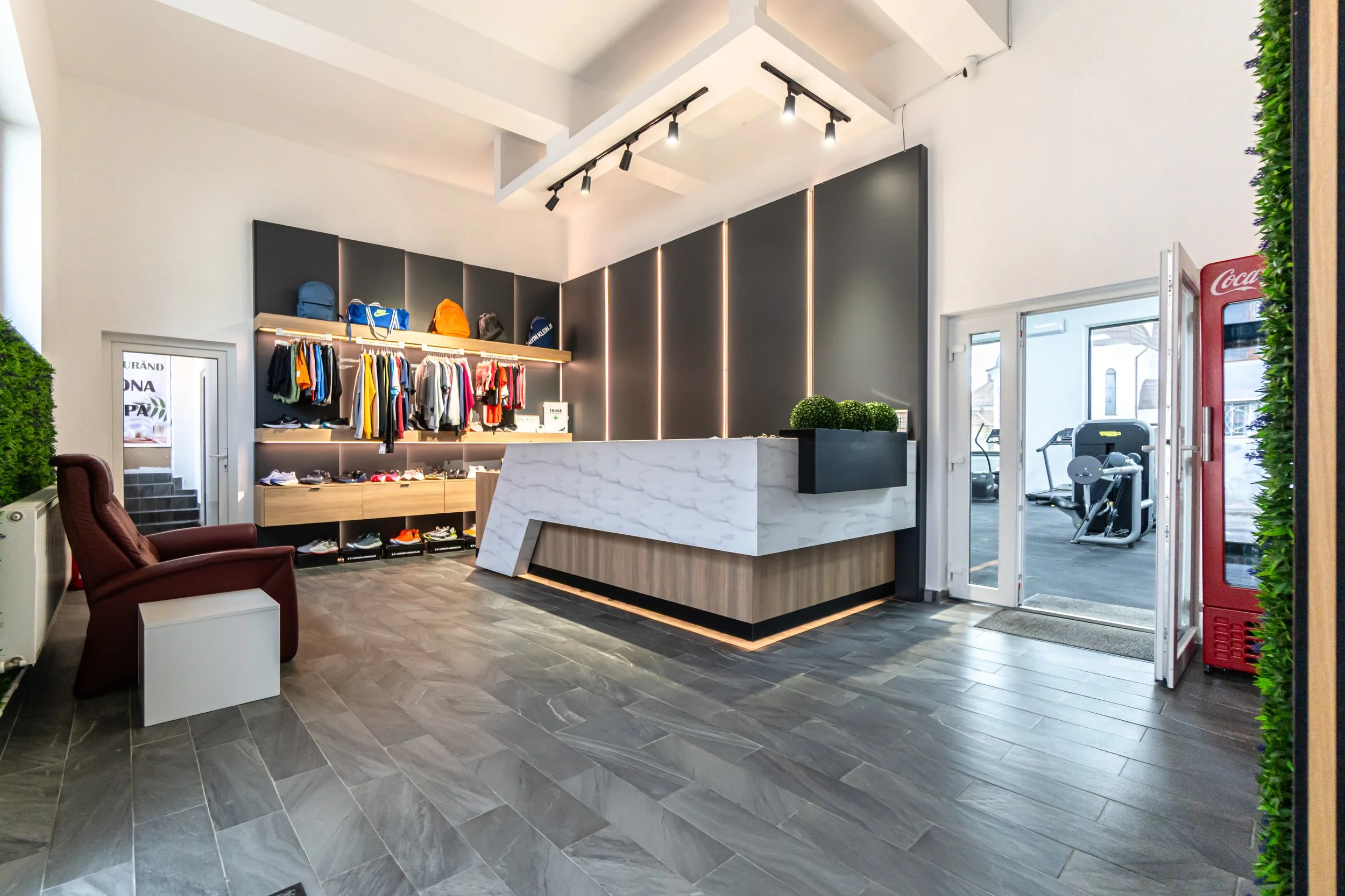 Modern retail store interior with a white marble checkout counter, retail clothing display with shirts and backpacks, a red Coca-Cola vending machine, and gym equipment visible through the glass door.