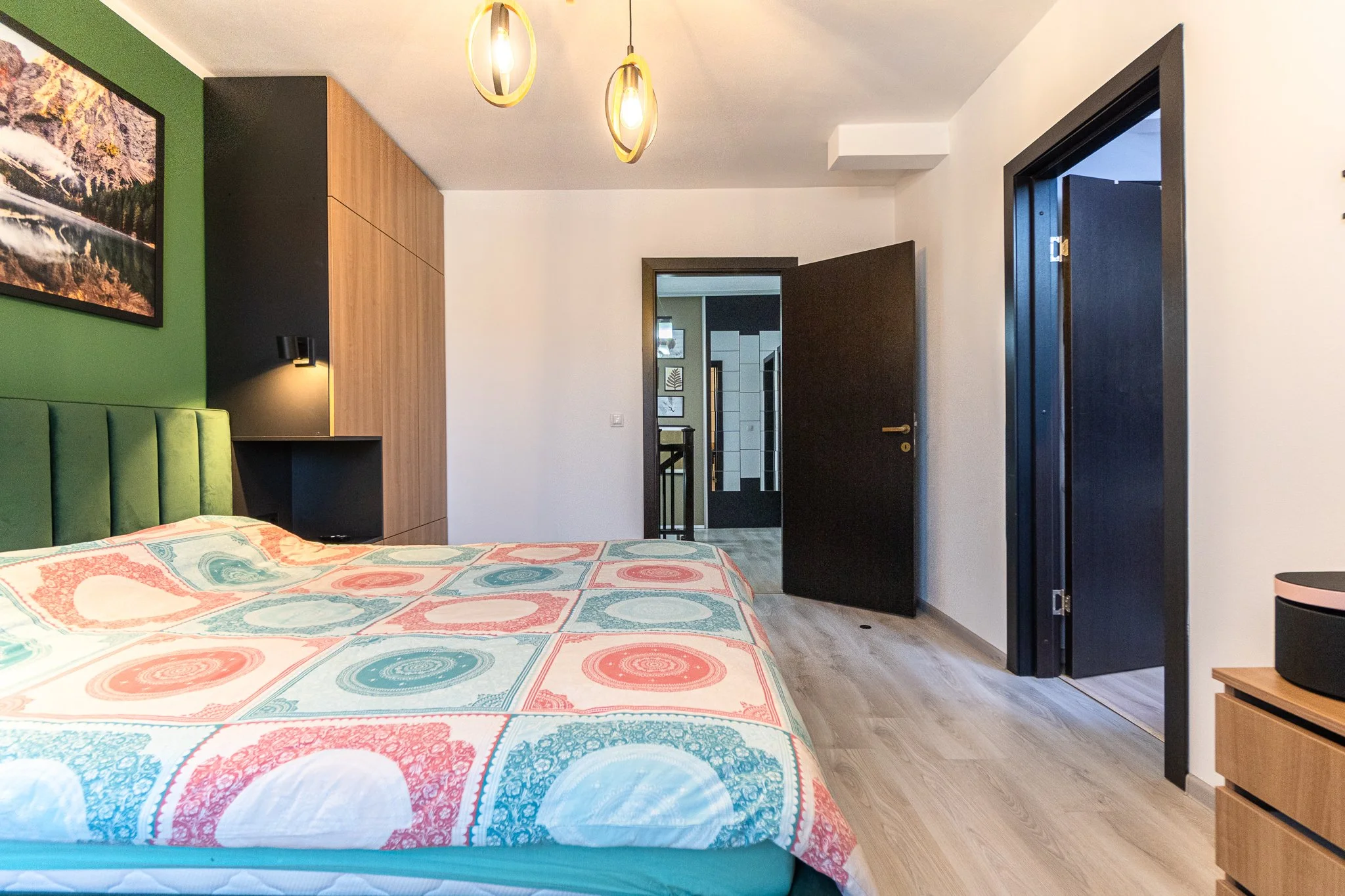 A bedroom with a green headboard and colorful patterned bedspread. Modern wooden and black closet doors, black door leading to another room, and wooden nightstand. Pendant light fixtures hanging from ceiling.