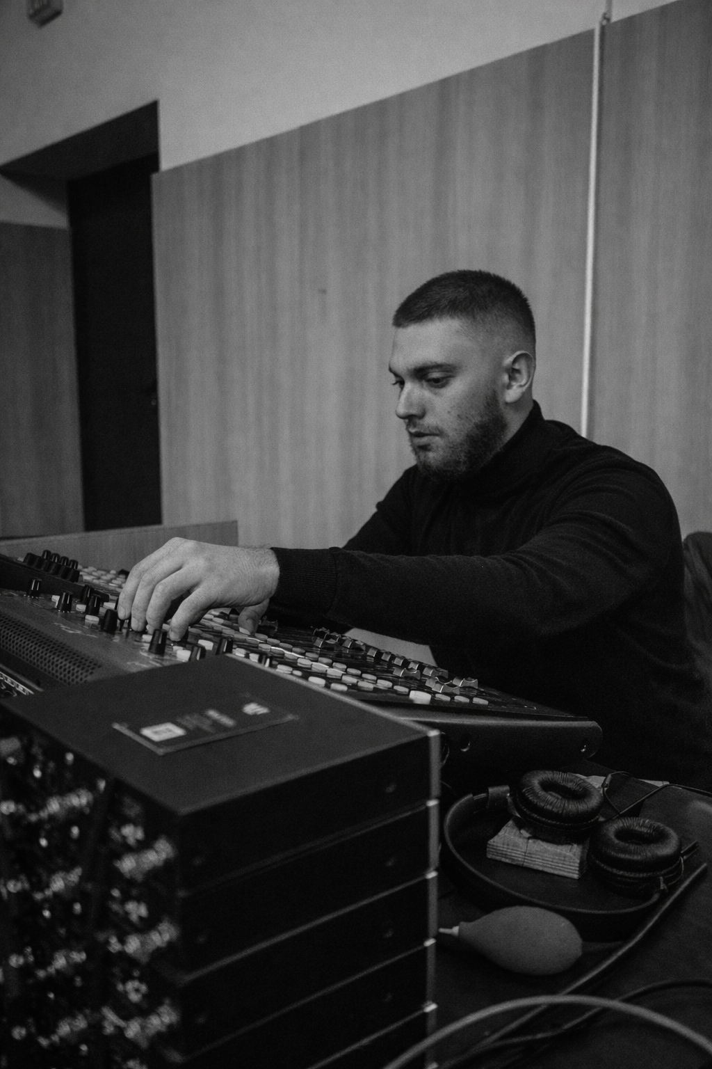 A man with a beard and short hair operating a DJ mixing console in a room with plain walls.