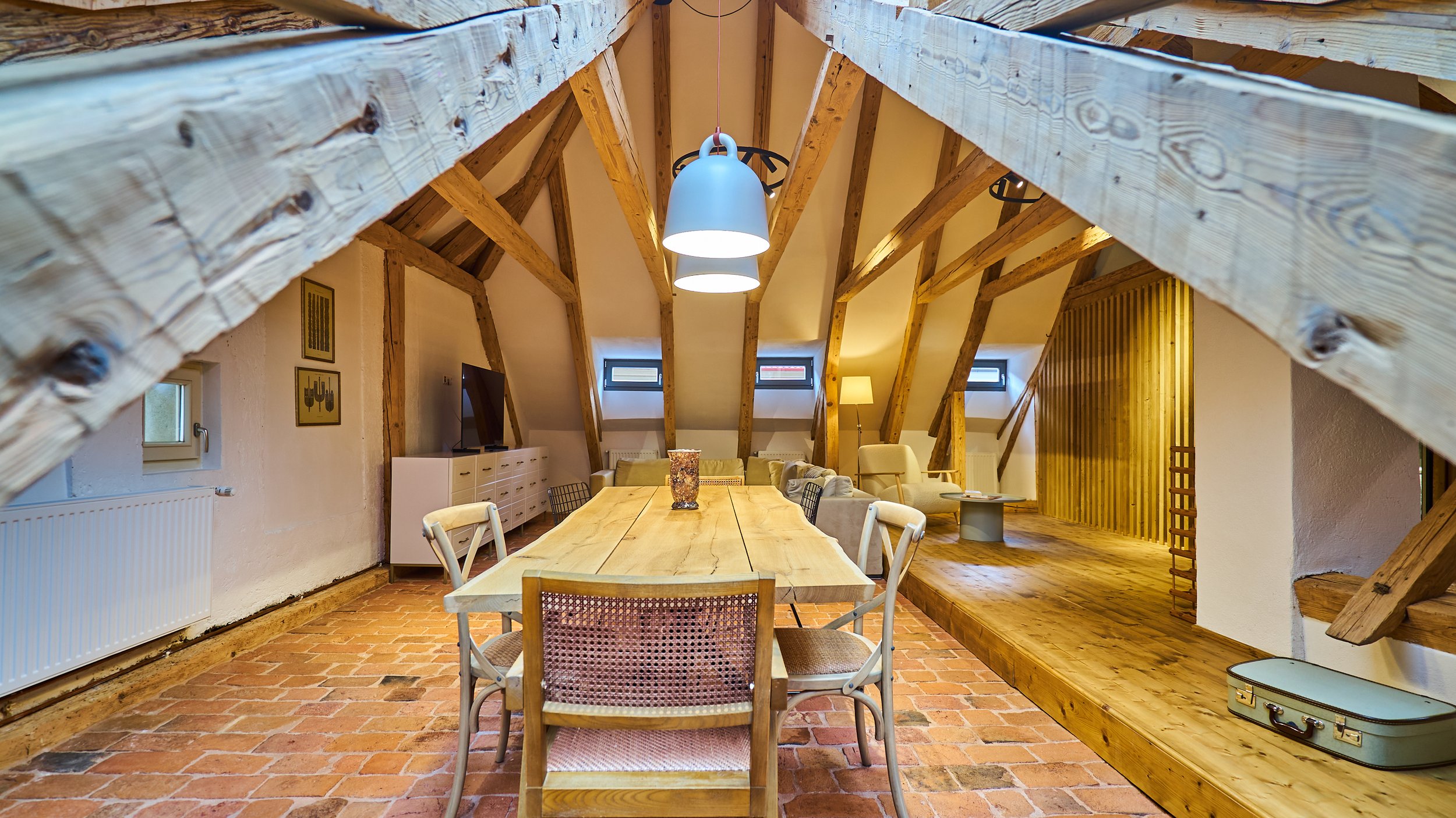 Interior of a cozy attic living space with sloped wooden beams, brick flooring, a dining table with chairs, and a sofa area with a television and floor lamp.