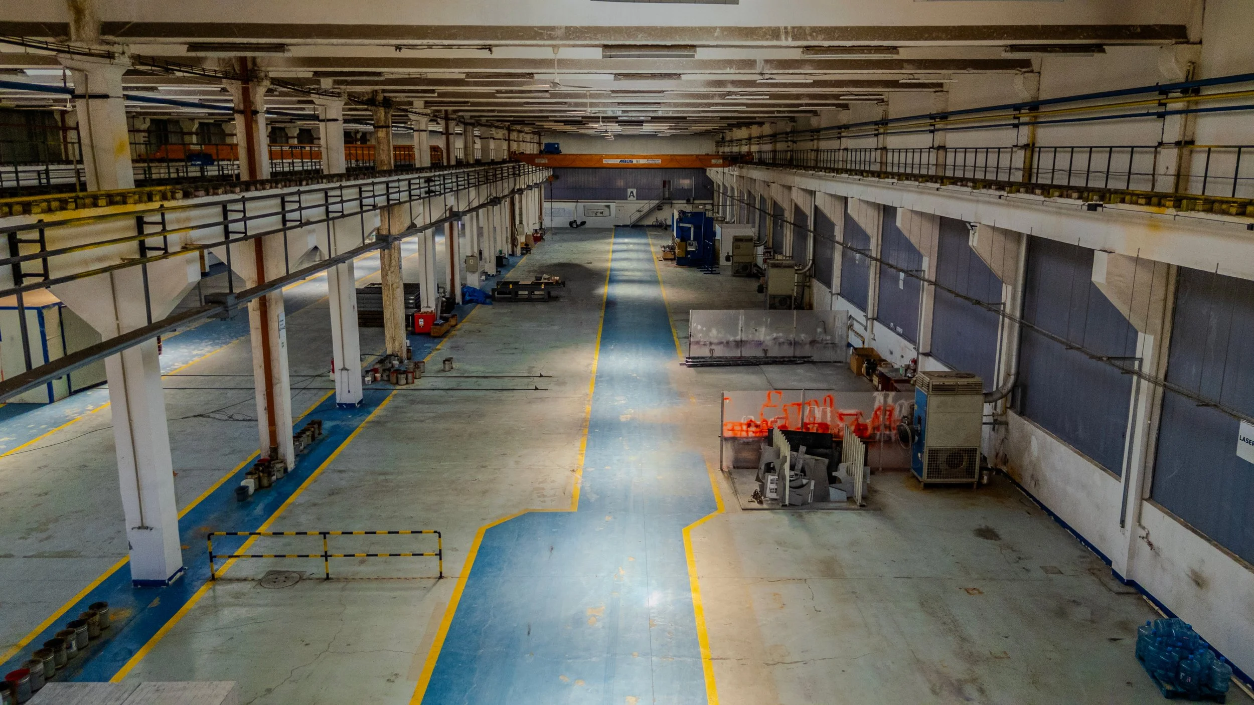 Empty industrial warehouse with yellow and blue painted lines on the concrete floor, storage equipment, and overhead crane.
