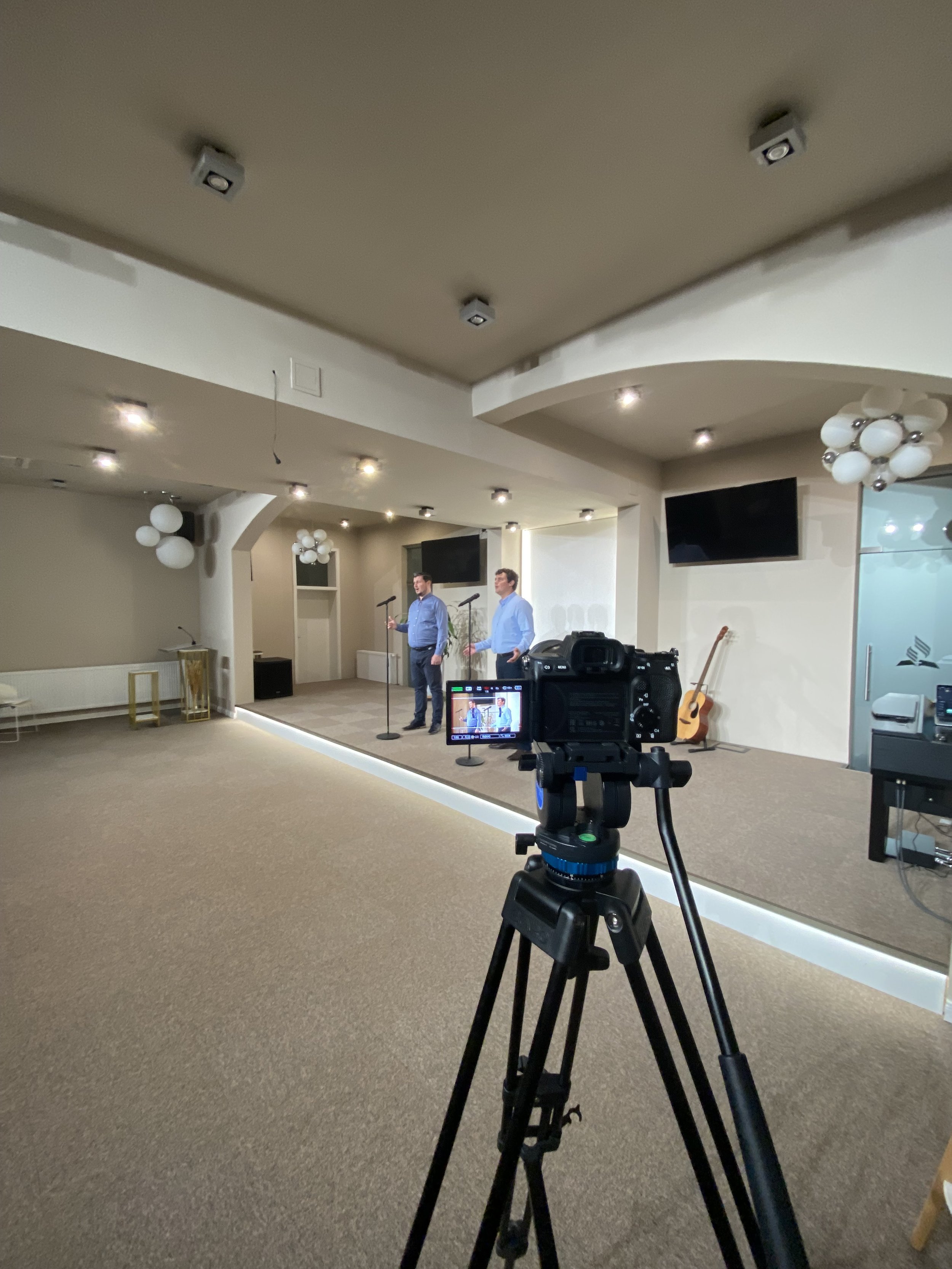 Two men speak on a stage with microphones, a guitar is leaning against the wall, and a camera is recording the scene in a conference room or event space.