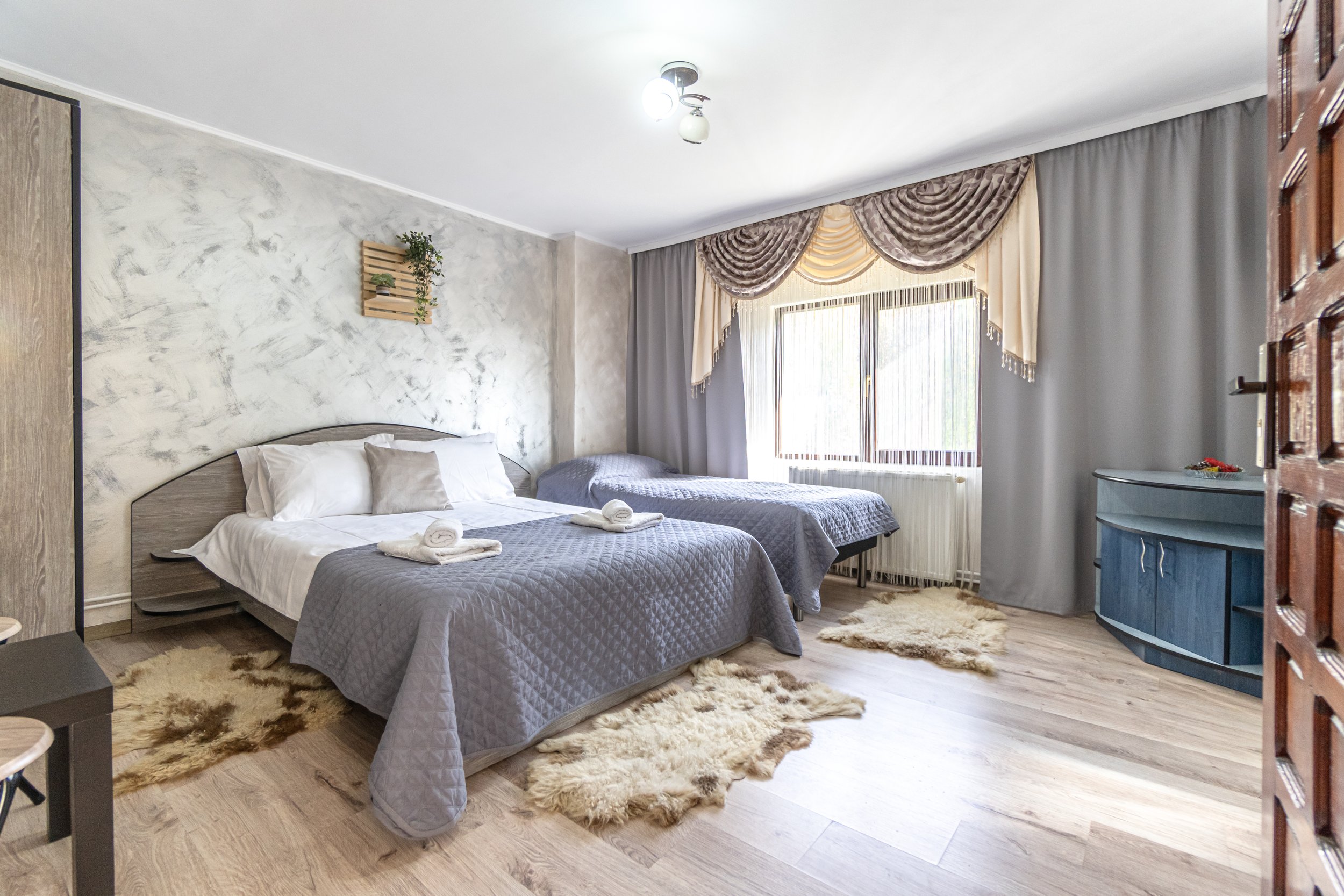 Hotel room with two beds, gray bedspreads, white pillows, and towels. Wooden wardrobe, blue dresser, window with curtains, and beige shag rugs.