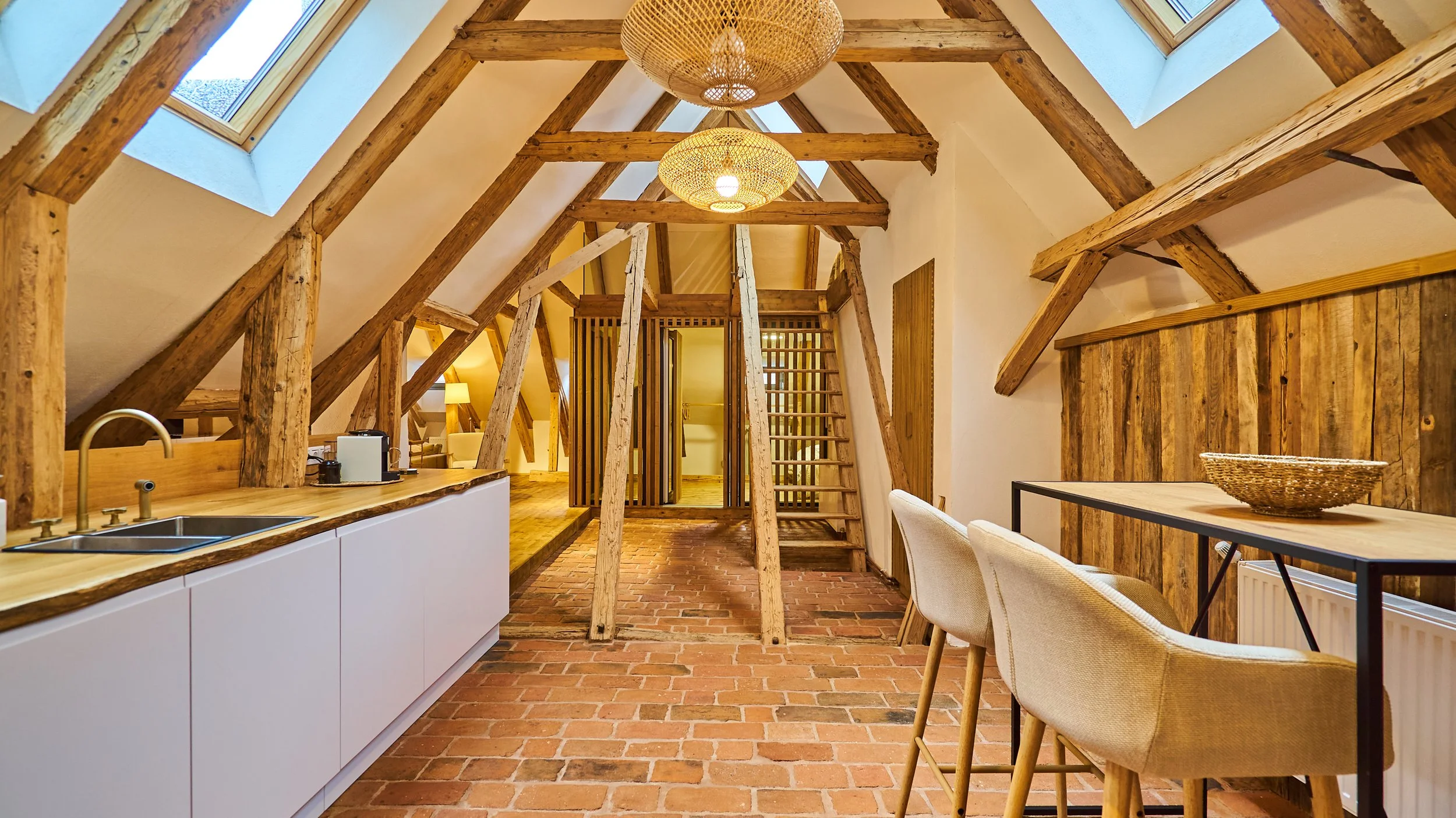 Interior view of an attic apartment with exposed wooden beams, brick flooring, skylight windows, and a small kitchenette with a sink, along with a dining area with chairs and a table.