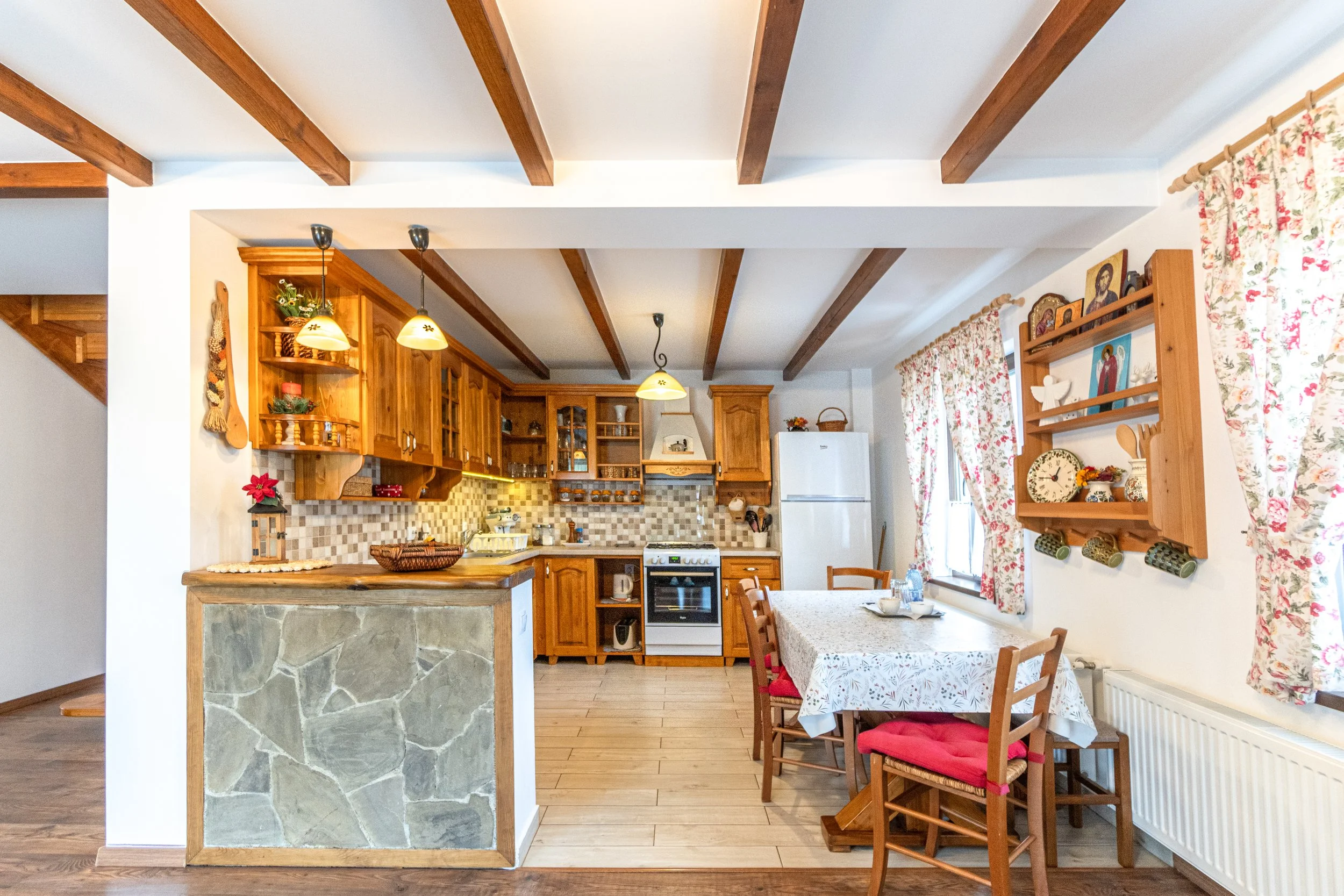 Cozy kitchen with wooden cabinets, a dining table with floral tablecloth and wooden chairs, floral curtains, and decorative shelves with dishes and ornaments