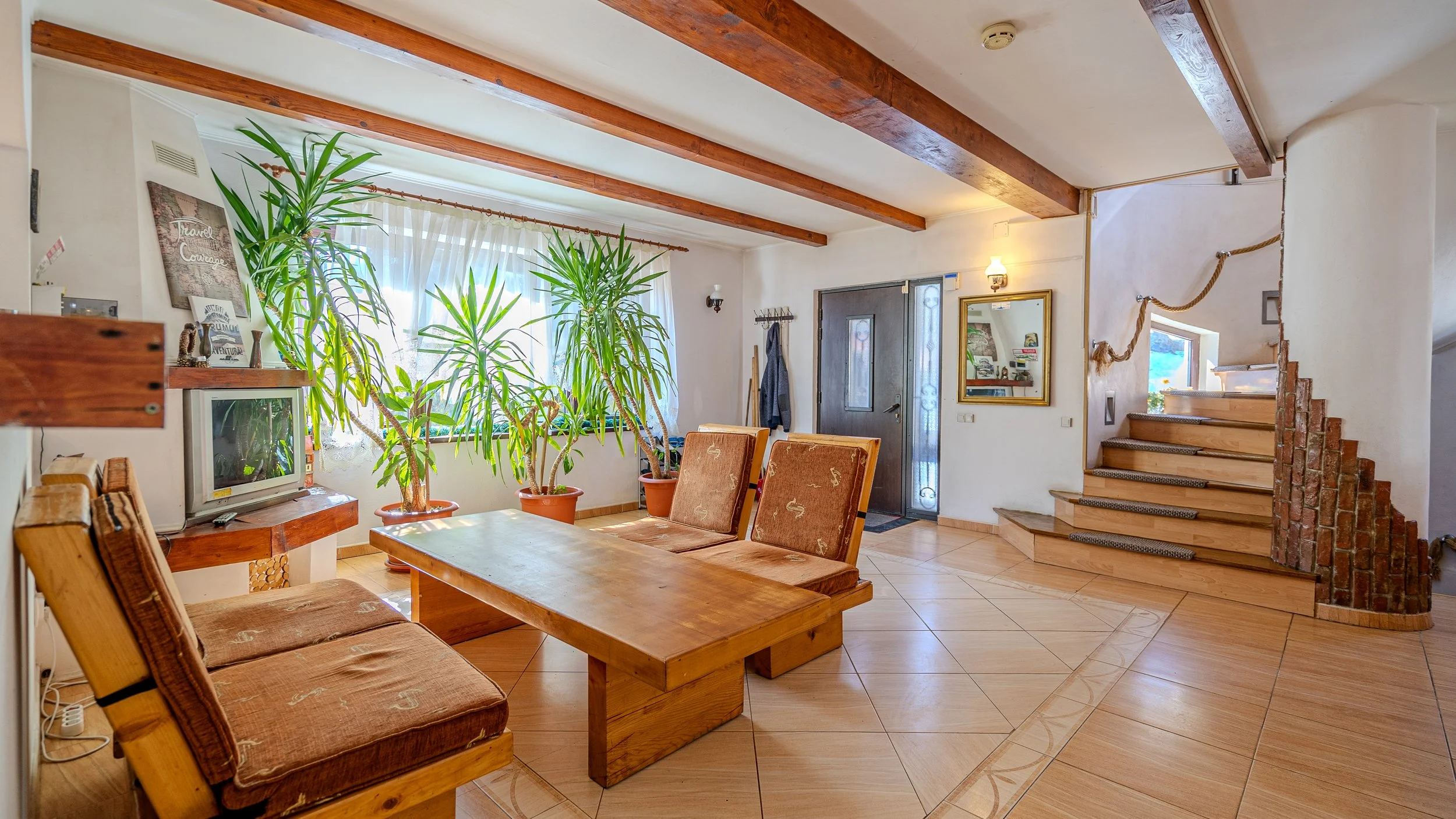 Living room with wooden beams, large potted plants, a wooden coffee table with cushioned chairs, a small television, a window with curtains, stairs with brick and wood design, and some decorative wall hangings.