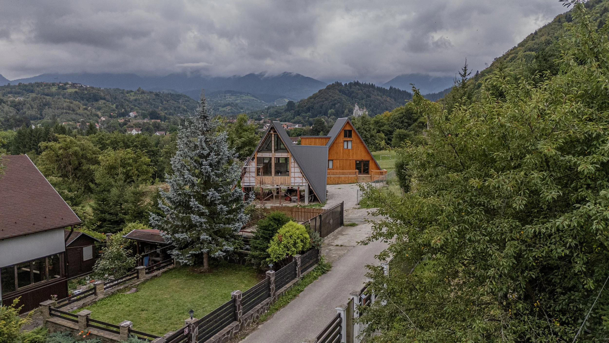 A scenic view of a hillside with two houses, one with a triangular roof with exposing wooden beams and the other with a brown roof, surrounded by lush green trees, and mountains in the background under a cloudy sky.
