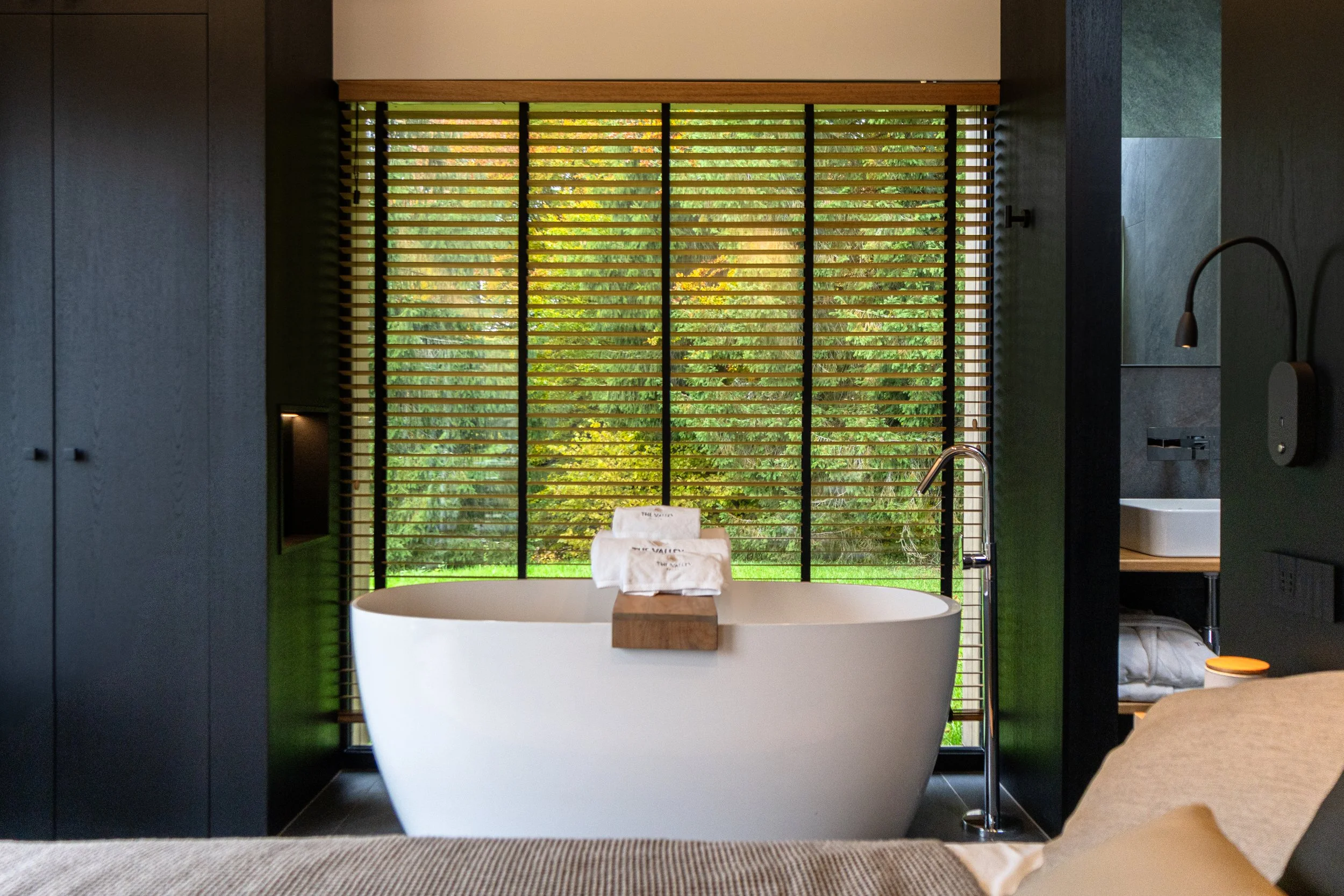Modern bathroom with freestanding white bathtub in front of large window with wooden blinds, overlooking green outdoor scenery, with black and dark gray walls and minimalist fixtures.