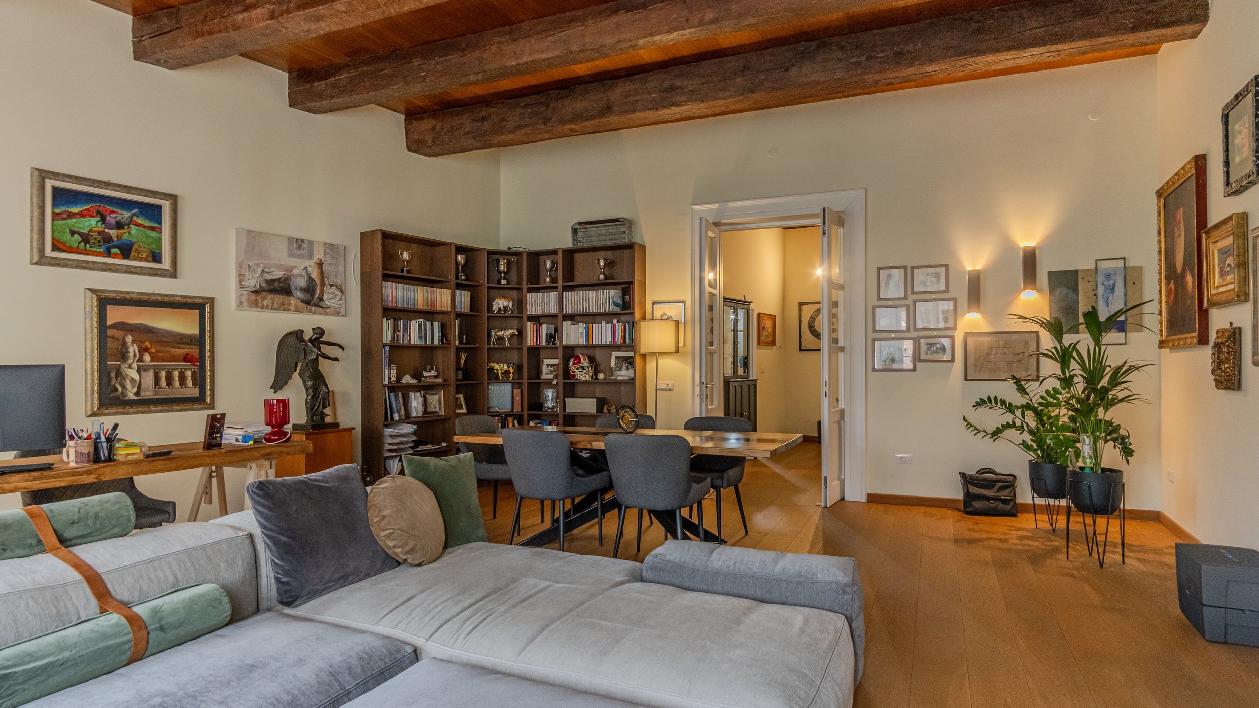 Living room with wooden floors, beige walls, exposed wooden beams ceiling, gray sofa with colorful pillows, wooden dining table with six chairs, large wooden bookshelf filled with books and decorative items, artwork on walls, houseplant in a black po