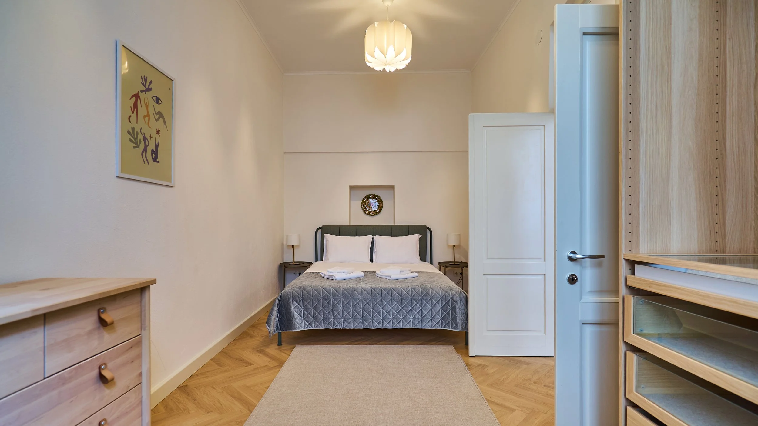 A minimalist bedroom with a double bed, white bedding, and two pillows. There are two small nightstands with lamps on either side of the bed, a decorative wall hanging above it, and a painting on the left wall. The room has wooden flooring and a ceil