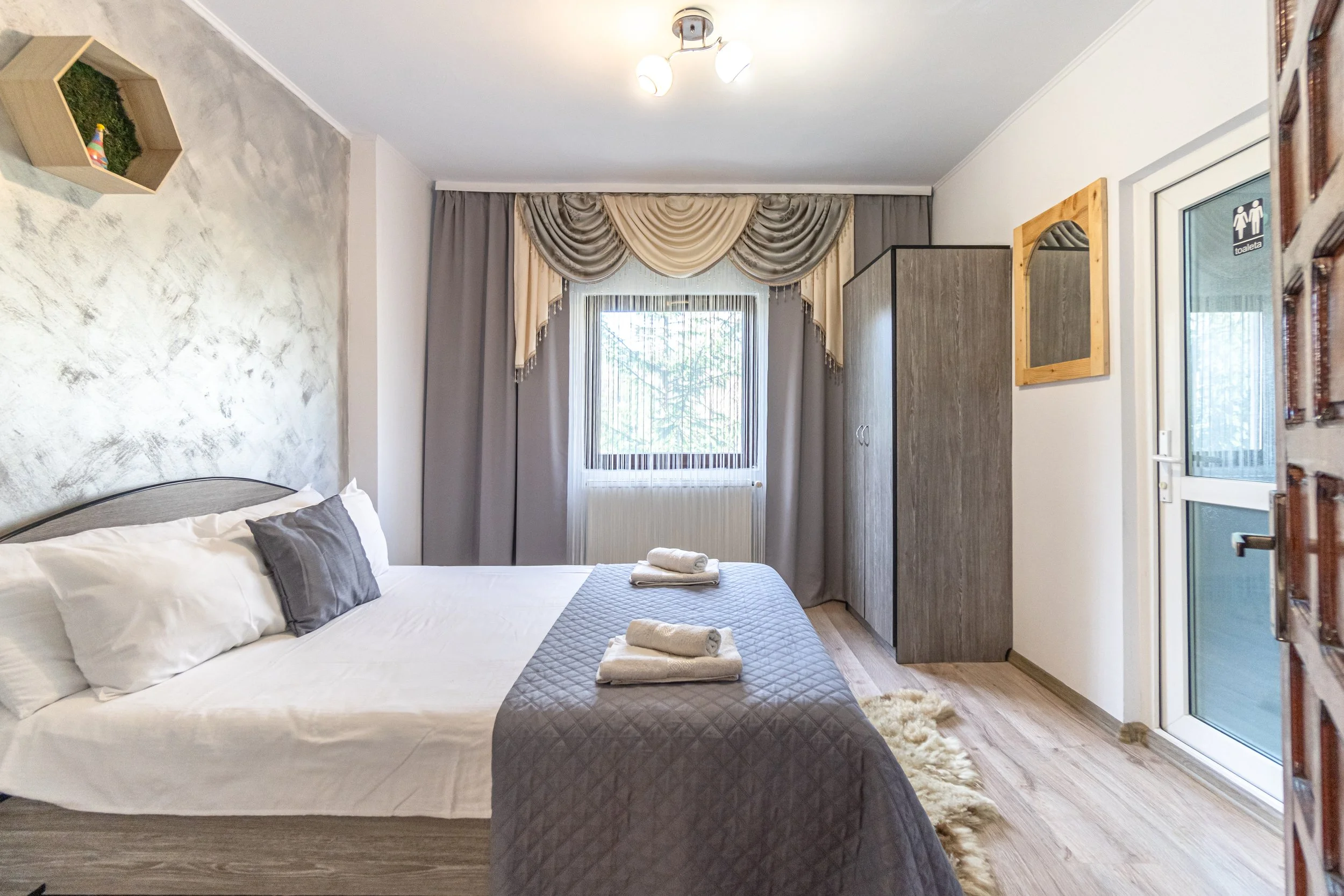 A bedroom with a bed covered with a gray quilt and white pillows, three folded towels on the bed, a window with drapes, a wardrobe, a framed mirror, a door with a restroom sign, and hardwood flooring.