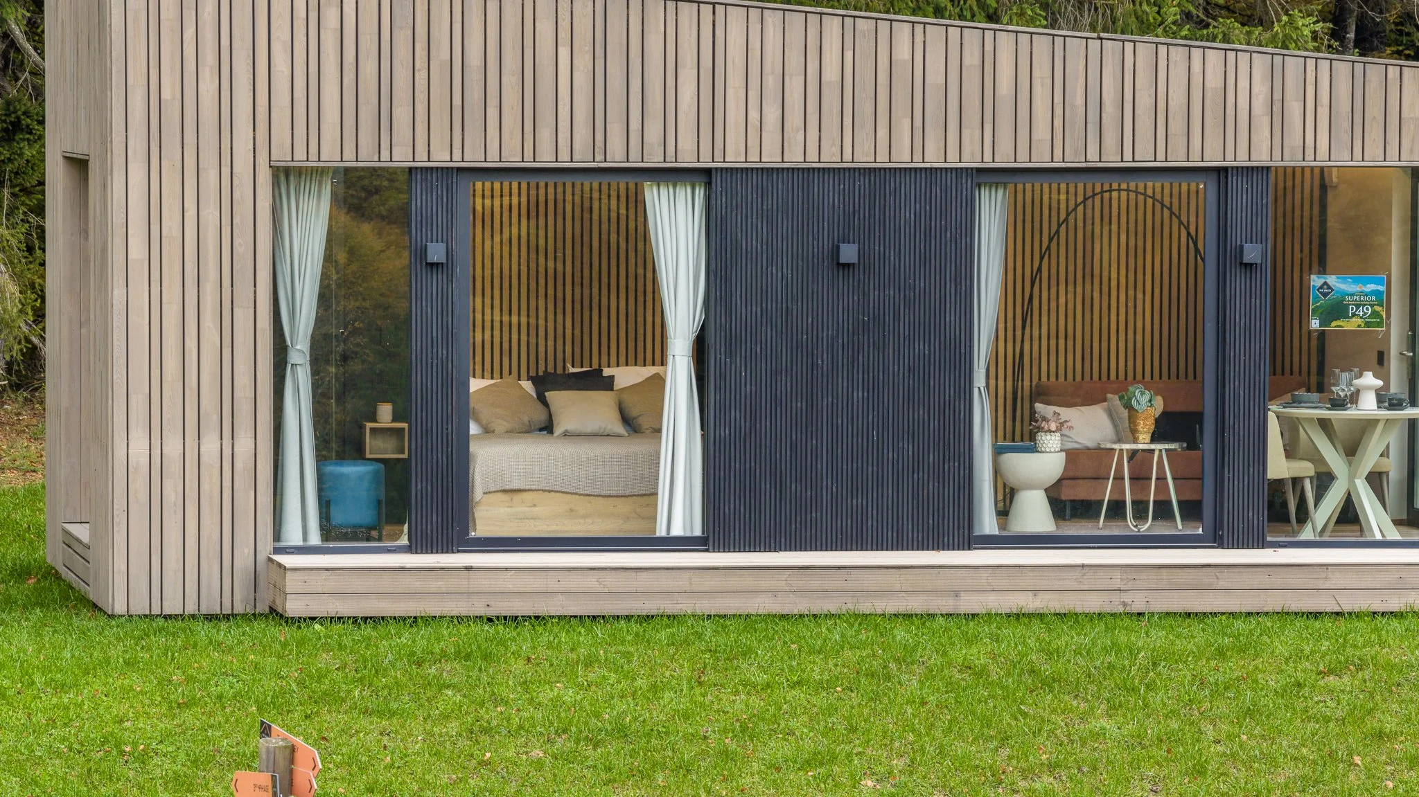 Modern tiny house with large glass windows, showcasing a bedroom with beige bedding and pillows on the left, and a living space with a brown sofa, side table, and dining area on the right. The house has wood panel exterior and is set on a grassy lawn