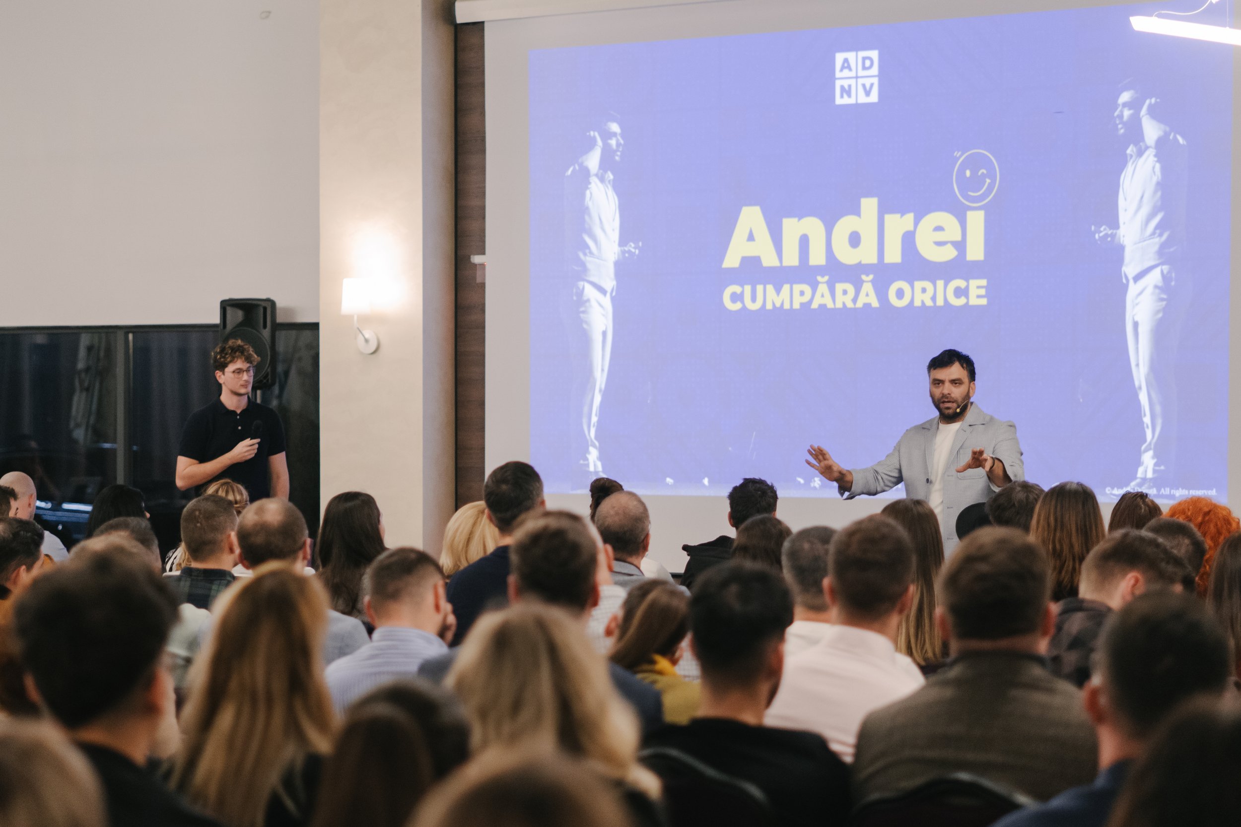 A man in a light gray blazer is speaking to an audience in a conference room, with a large screen behind him displaying the text 'Andrei Cumpără Orice' and a logo. A young man holding a microphone is standing on the side.