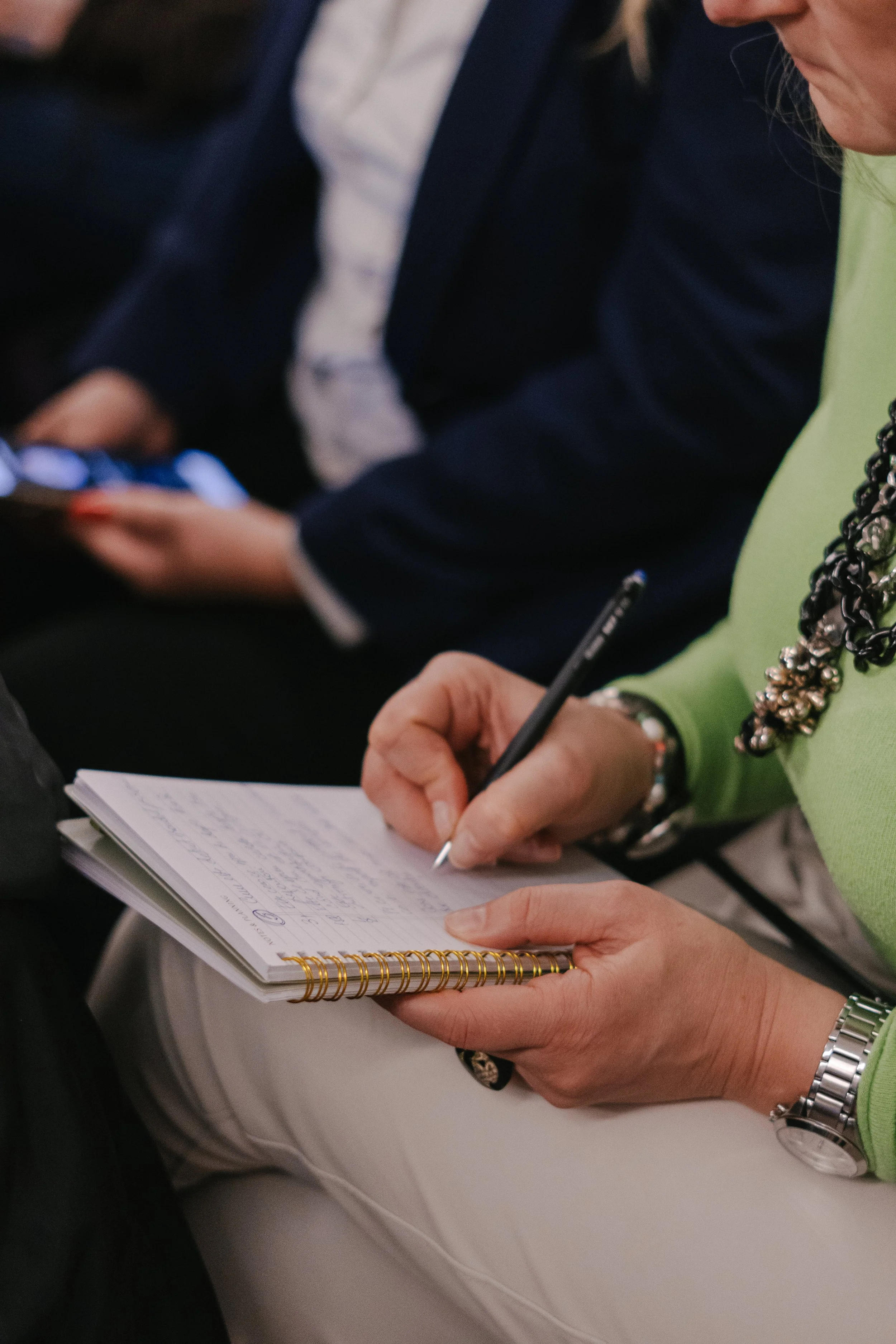A person in a green top and silver watch is writing in a notebook with a black pen, holding the notebook with their left hand. Other people are sitting nearby, one wearing a blue blazer and a white shirt.