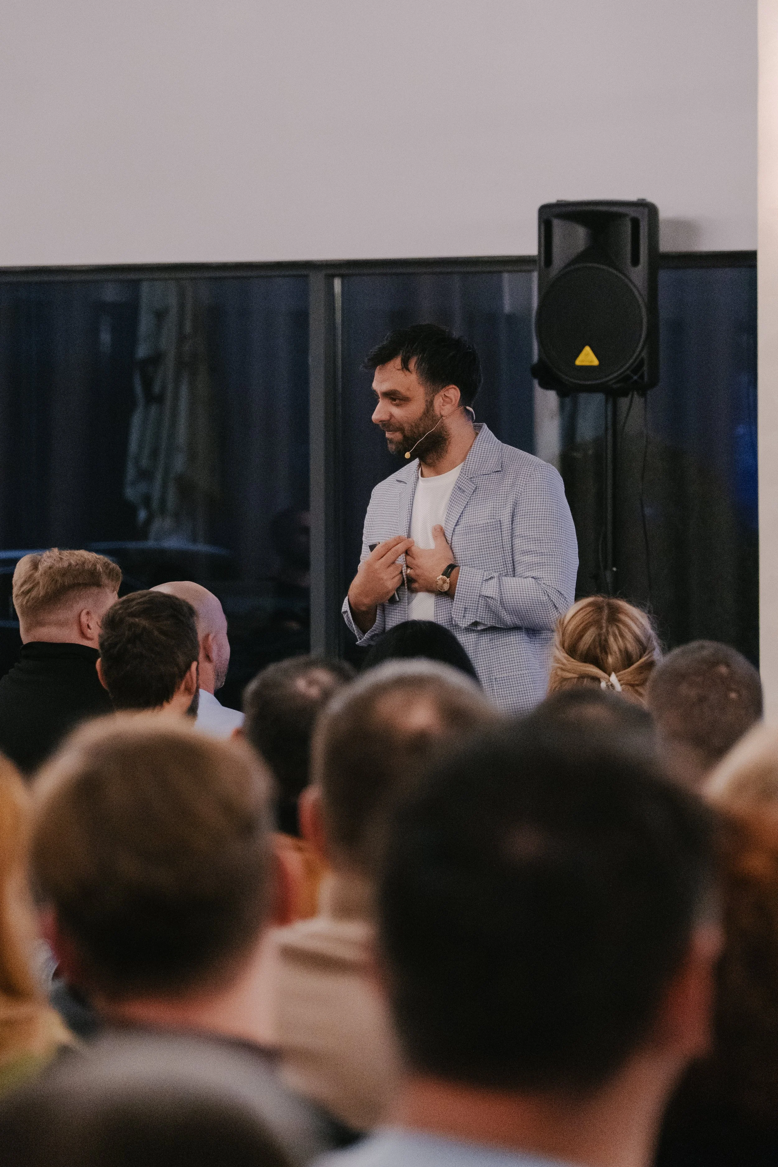 A man with dark hair and beard, wearing a light checkered blazer and a white shirt, stands in front of an audience, speaking or presenting. He has a microphone headset and is gesturing with one hand on his chest. There is a large speaker behind him a