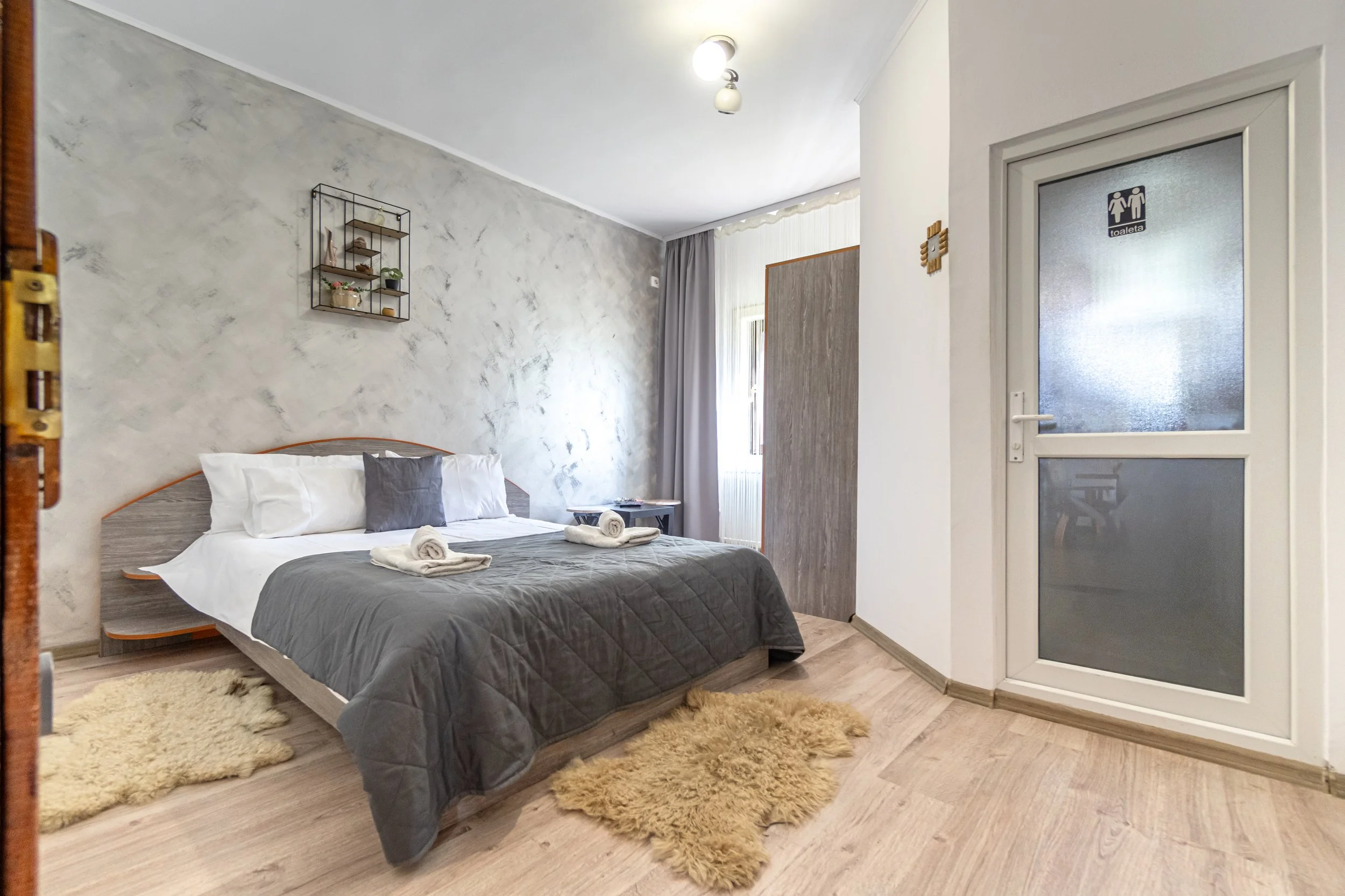 A cozy bedroom with a large bed dressed in white pillows and a gray bedspread, featuring two rolled towels on top. The room has wooden flooring, two beige rugs, a wall-mounted shelf with decorative items, and a window with curtains allowing natural l