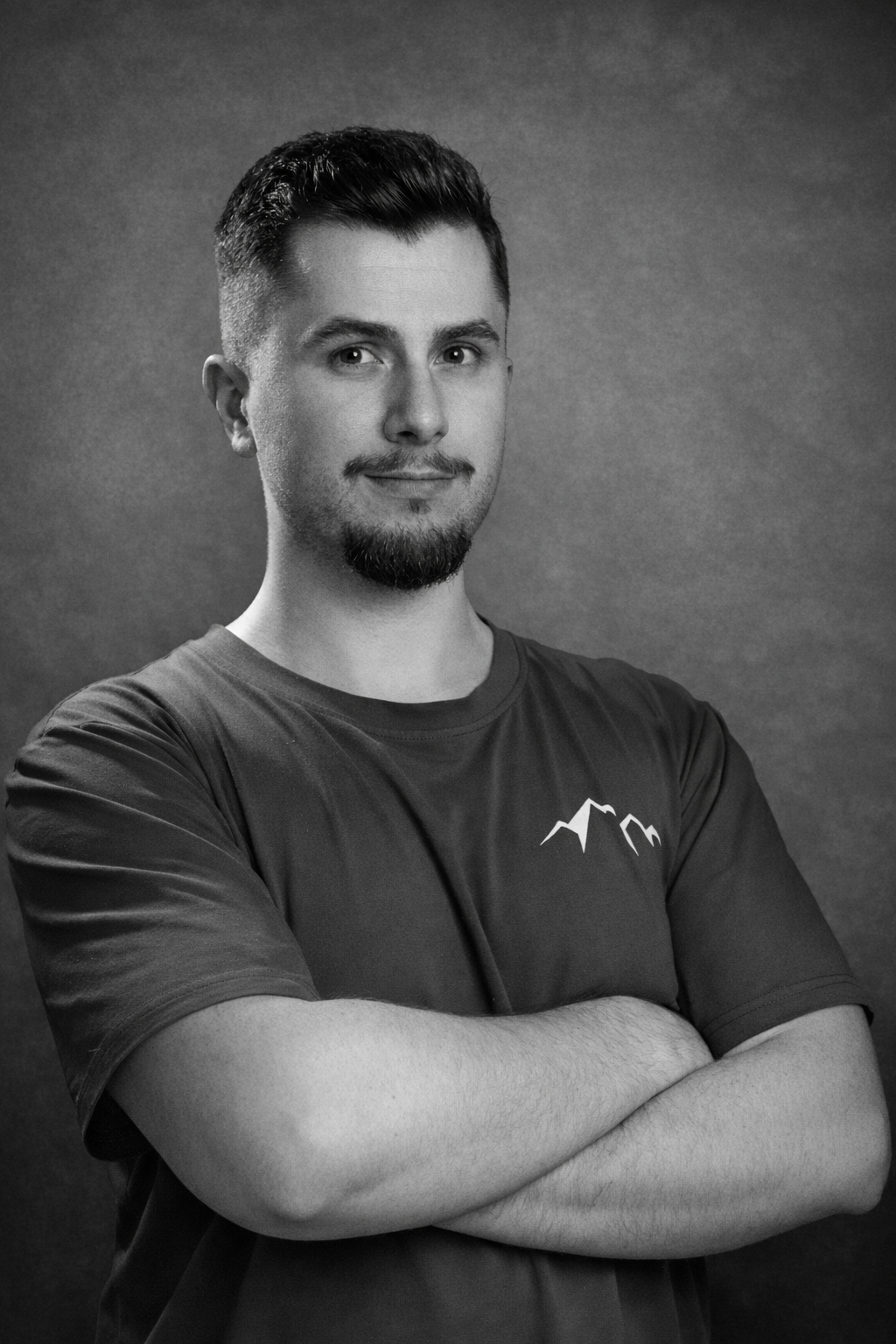 Black and white portrait of a young man with short dark hair and a beard, wearing a T-shirt with a mountain graphic, standing with arms crossed against a plain background.