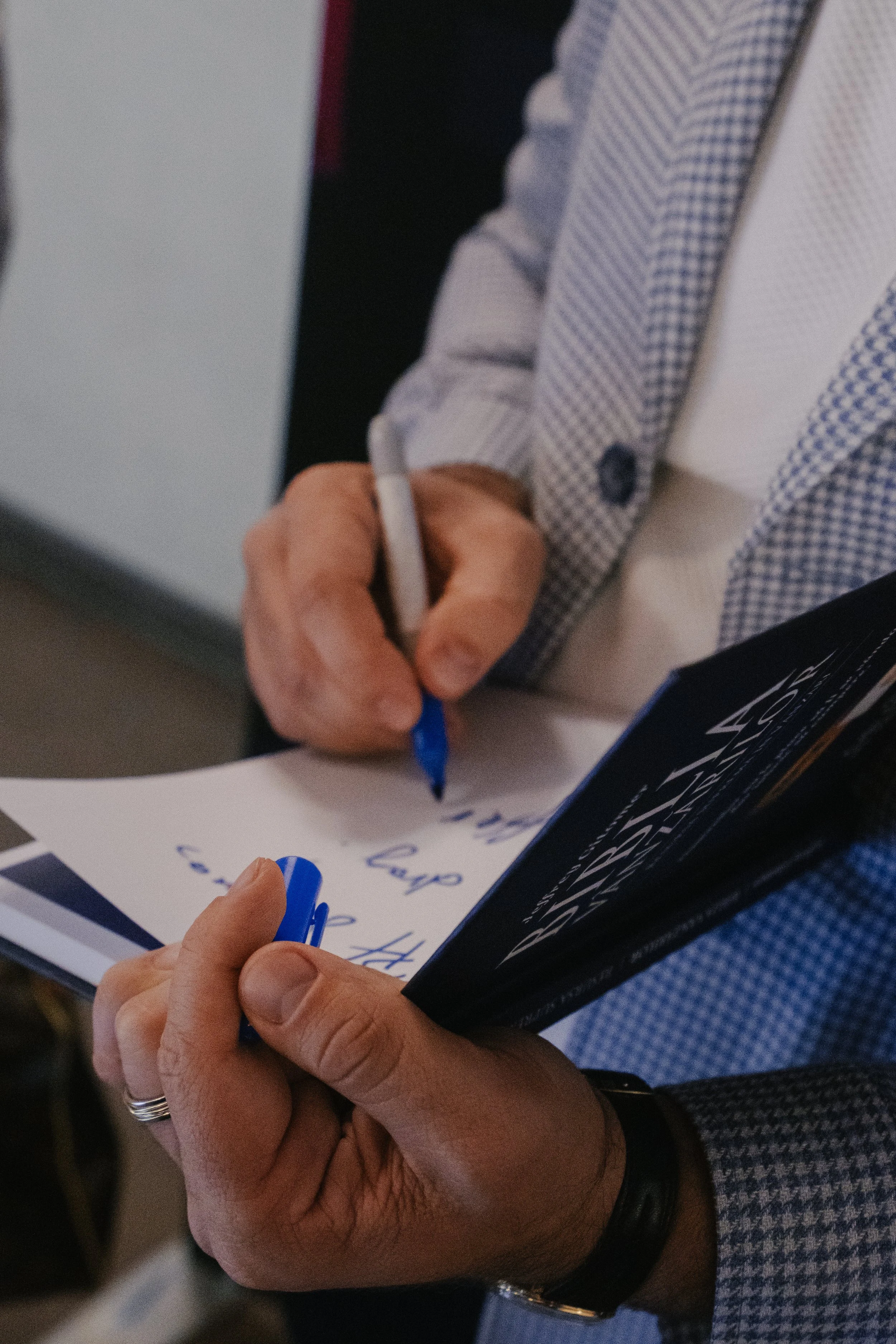 A person in a checkered blazer writing on notes with a blue marker, holding a black book or notebook.