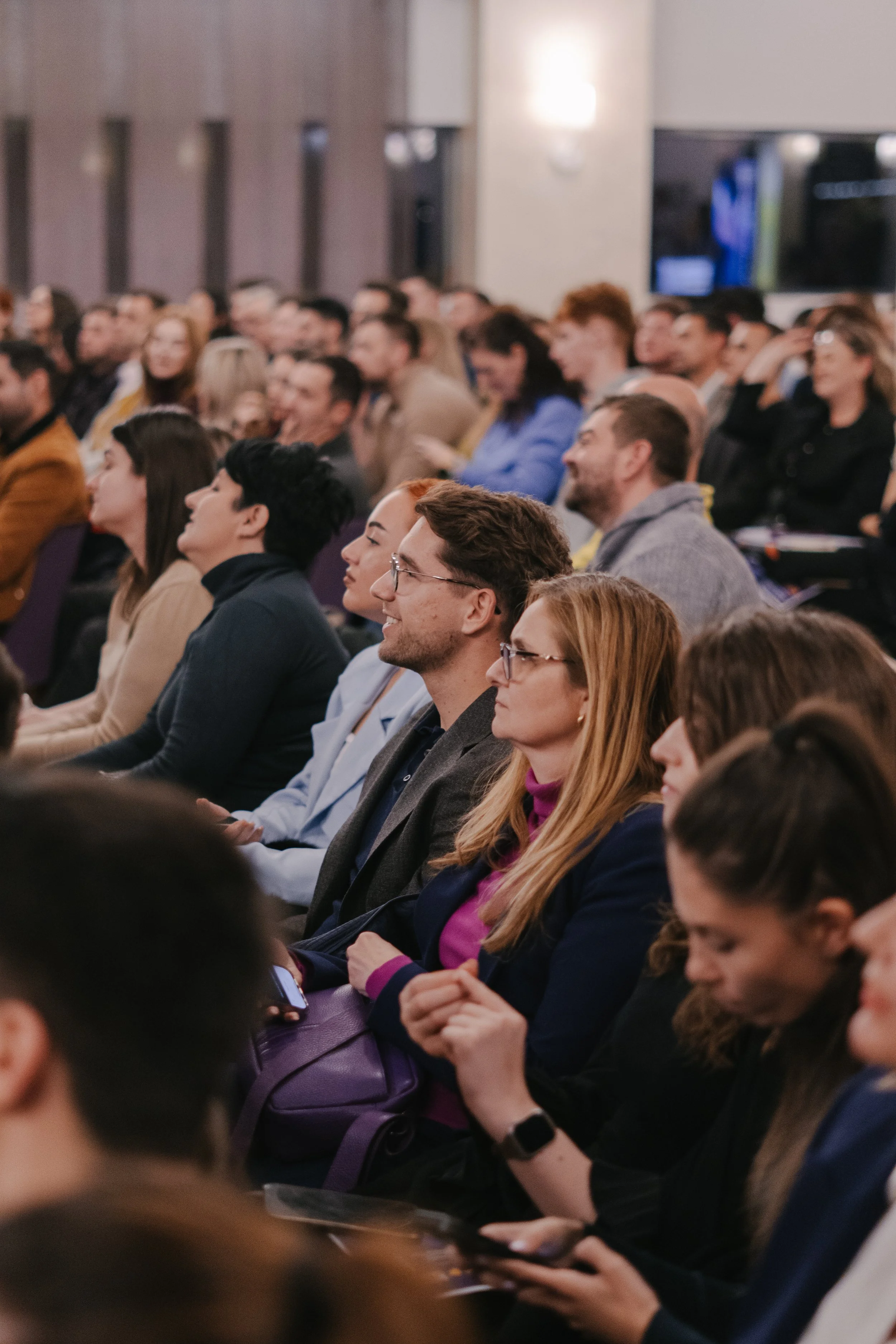 Audience attending a conference or seminar, focused and listening attentively in a large, well-lit room.
