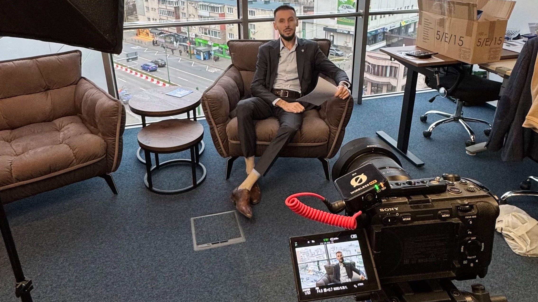 Man in a suit sitting on a brown armchair during a video interview in a modern office with large windows showing a city street. Camera equipment and a monitor are visible in the foreground.