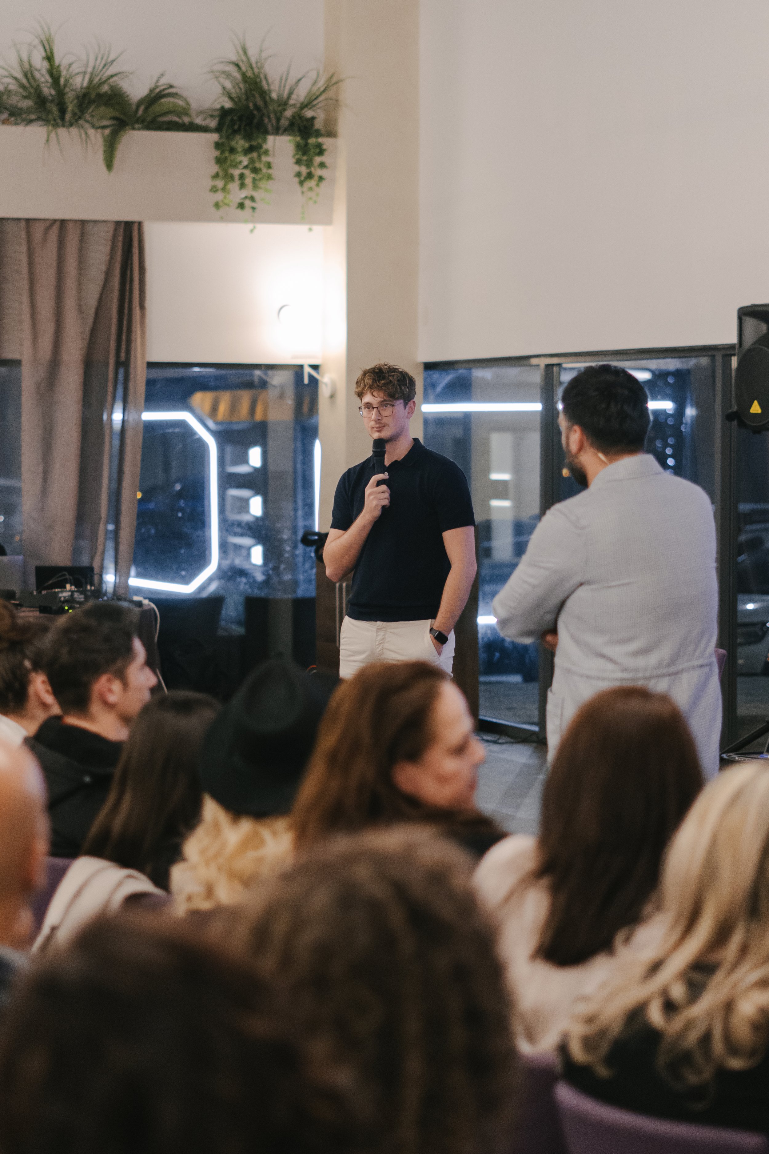 Young man with glasses speaking into a microphone at a presentation or event, with an audience of diverse adults seated and listening indoors.