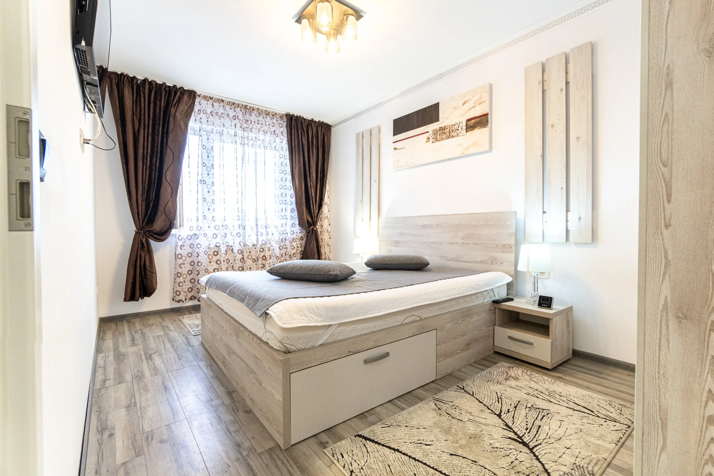 A bedroom with a bed, two pillows, nightstands, a window with brown curtains, and a piece of abstract wall art.