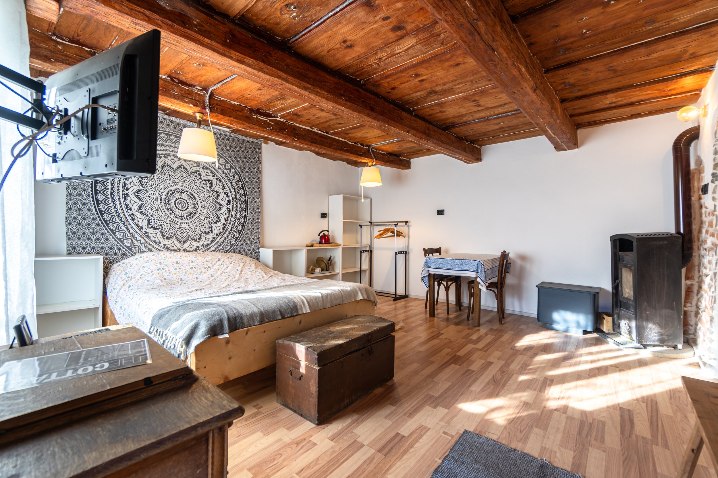 A rustic bedroom with a wooden ceiling, a bed with a floral blanket, a tapestry wall hanging, and a small dining area with a table and chairs near a wood stove.
