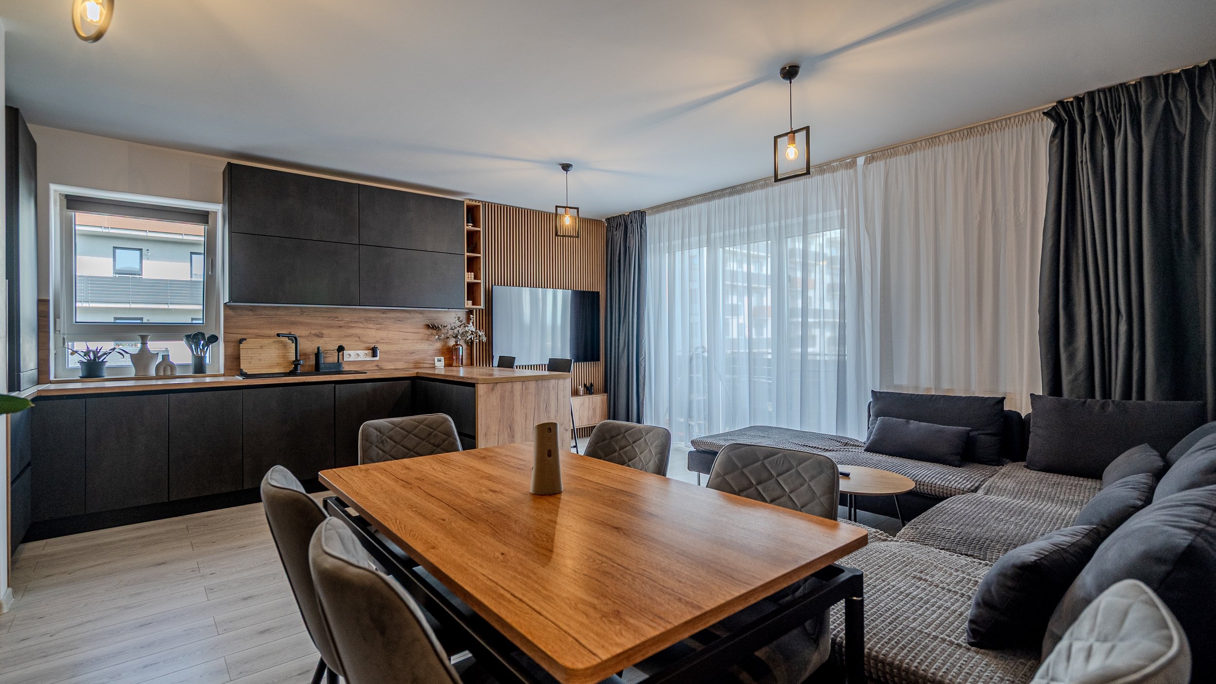 Modern living room and kitchen area with a wooden dining table, gray upholstered chairs, a gray sectional sofa, floor-to-ceiling curtains, and natural light from large windows.