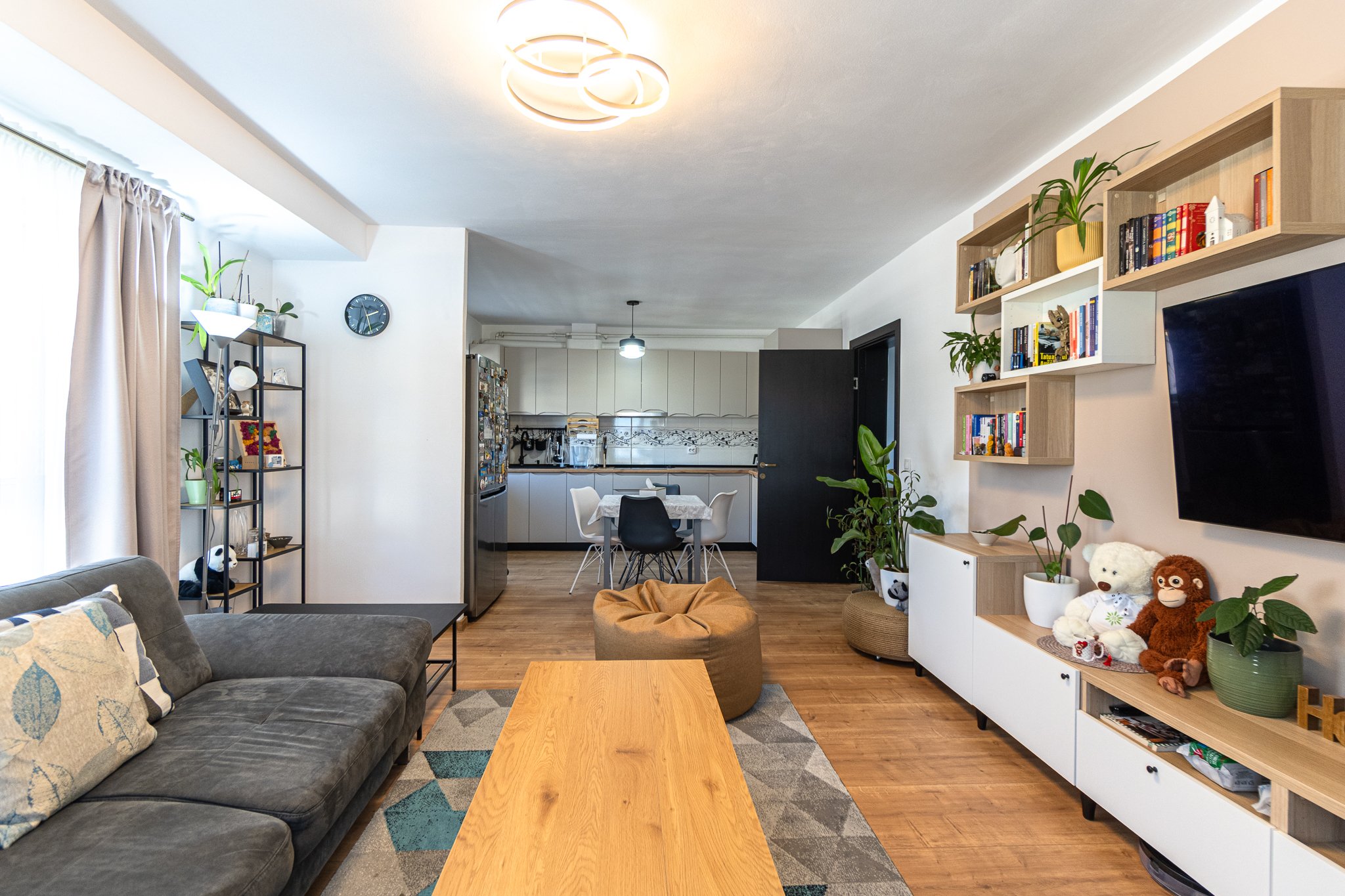 A living room with a gray couch, a wooden coffee table, bookshelf and TV on the right, and an open kitchen and dining area in the background.