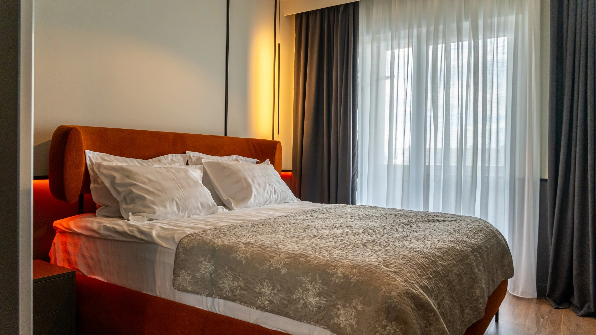 A hotel room with a made bed, white pillows, an orange headboard, and dark curtains with sheer curtains behind. There is a nightstand next to the bed.