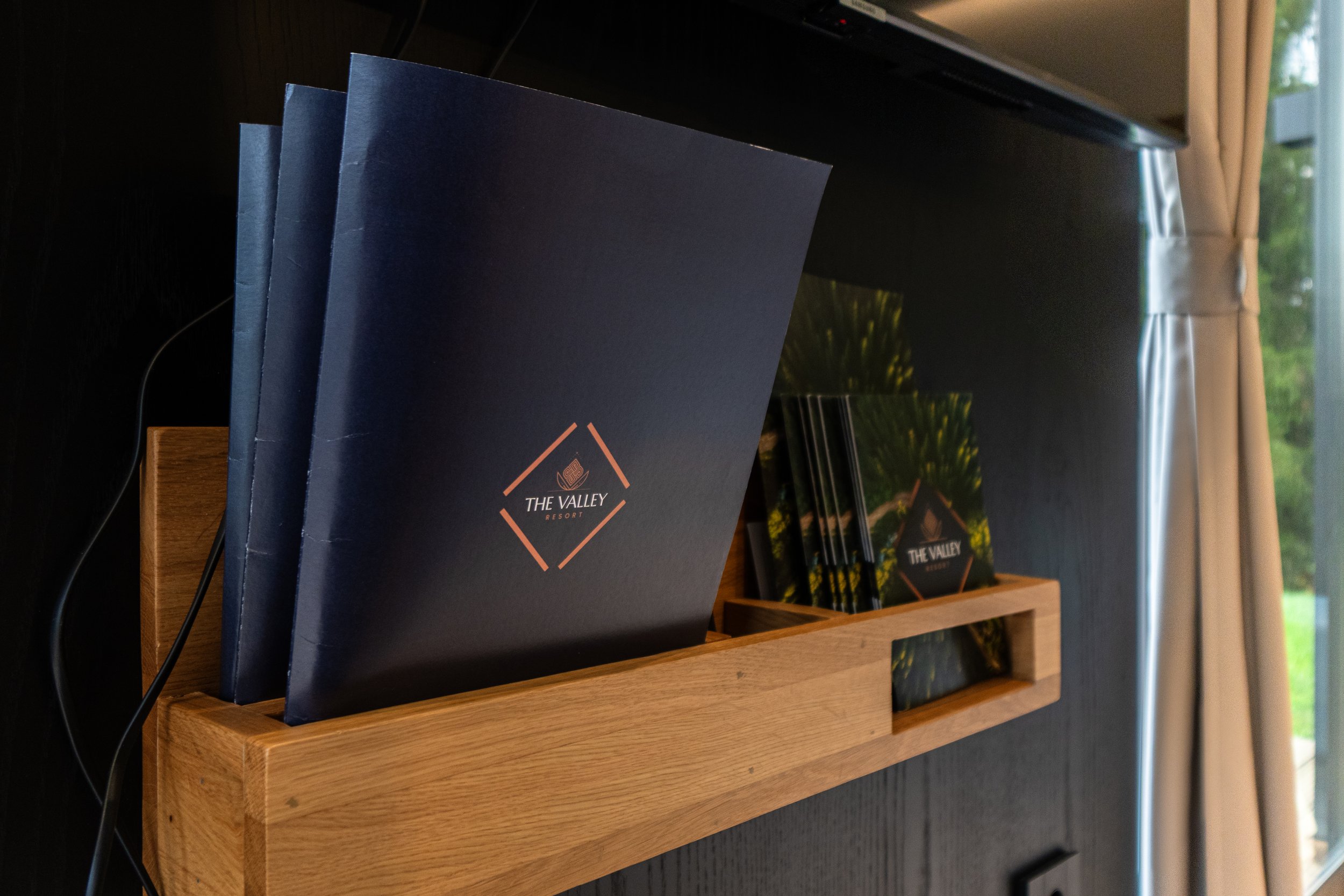 Promotional brochures and flyers for The Valley Resort on a wooden tray on a dark wall, with a window and curtains in the background.
