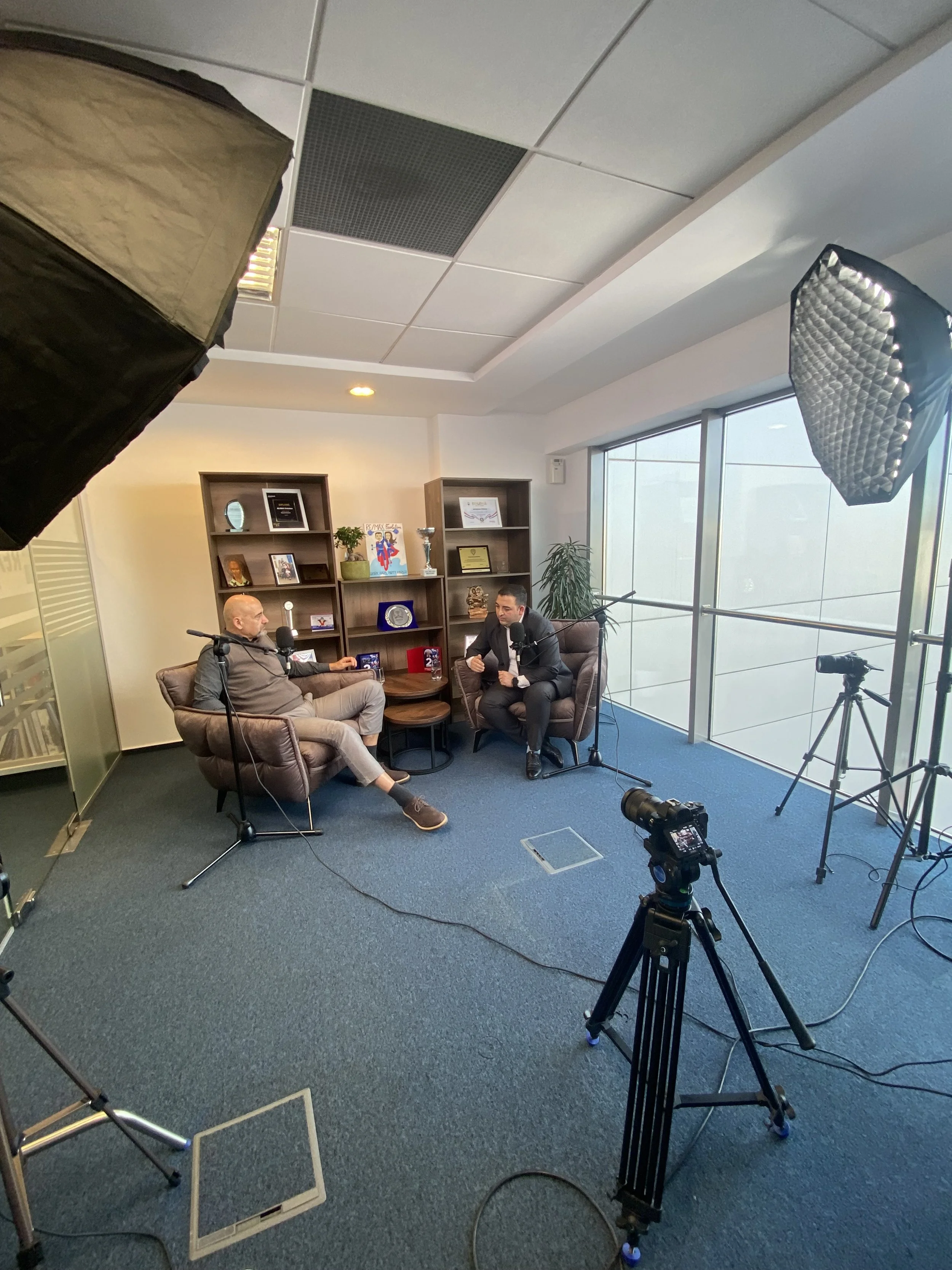 Two men are sitting in a recording studio, engaging in an interview. They are surrounded by professional camera equipment, including lights and tripods. The room features large windows, bookshelves with awards and framed pictures, and a small table between them.