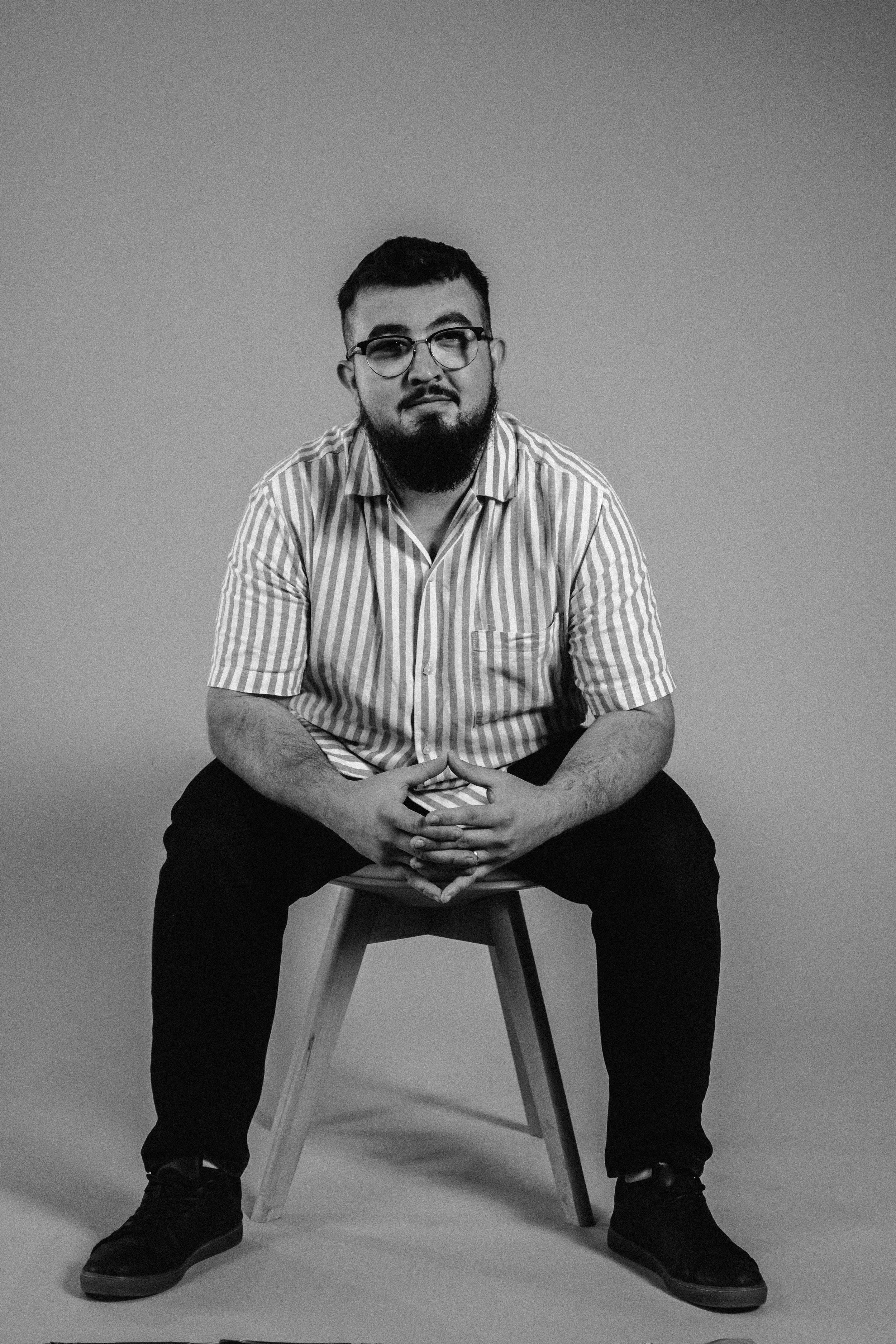 A man with glasses and a beard sitting on a wooden stool in a studio with a plain background, wearing a striped short-sleeve shirt and dark pants.