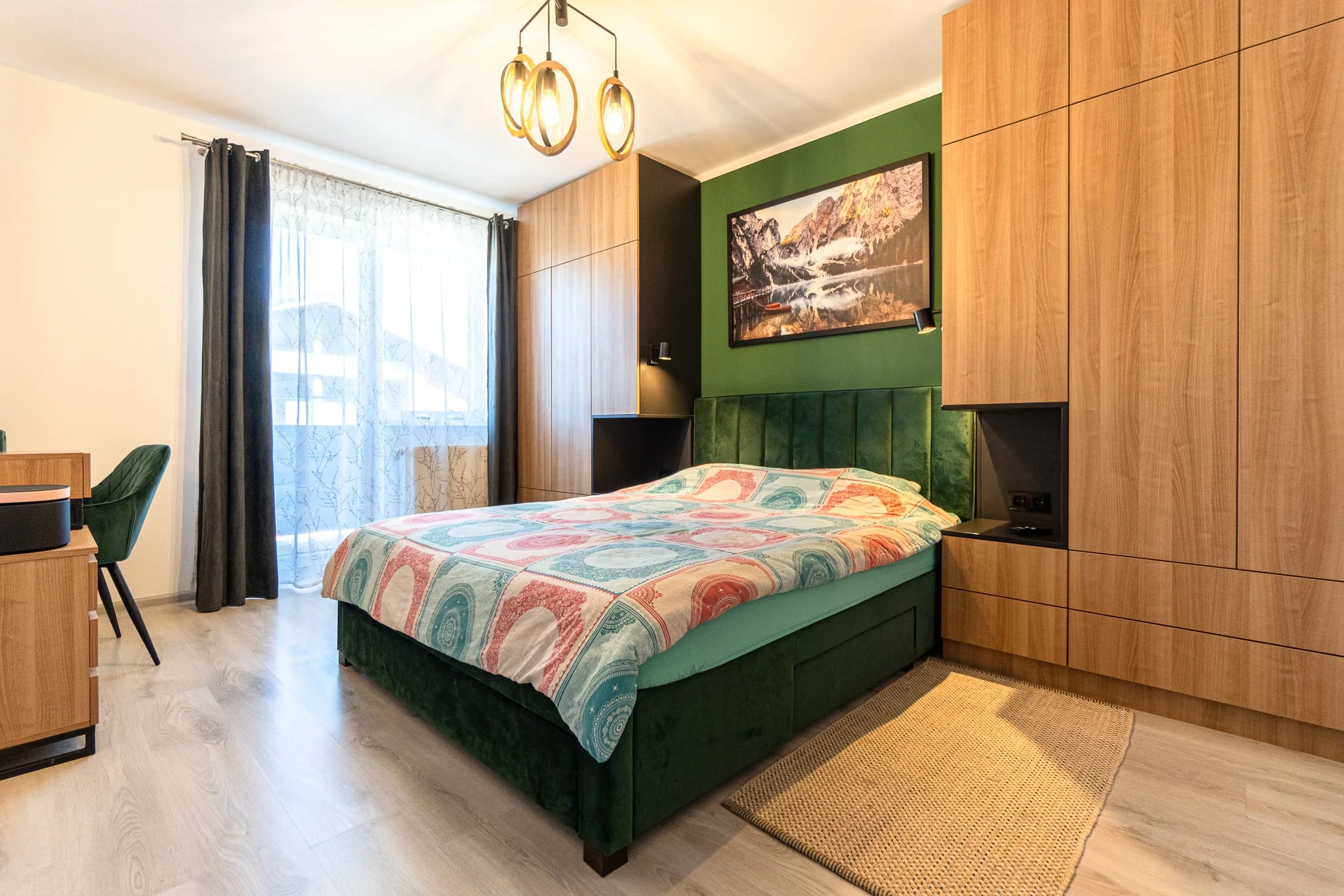 A modern bedroom with a green upholstered bed, colorful bedspread, wooden accent walls, and a framed landscape photo above the bed. There are black and wooden furnishings, a window with sheer and dark curtains, and a beige rug on light wood flooring.