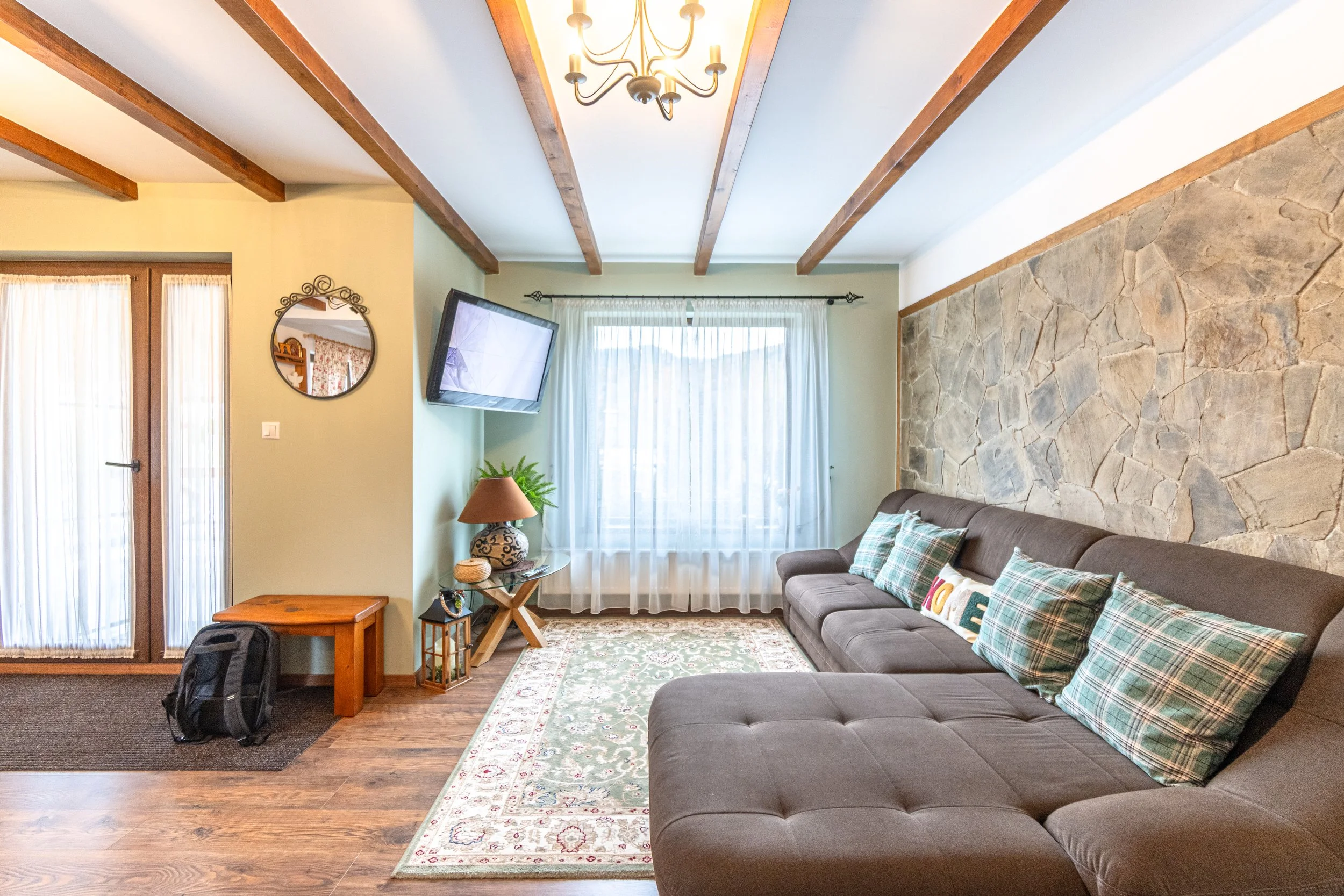 A living room with a brown sectional sofa with plaid and decorative pillows, a stone accent wall, a window with sheer curtains, a mounted flat-screen TV, a side table with a lamp, and a chandelier on a wooden-beamed ceiling.