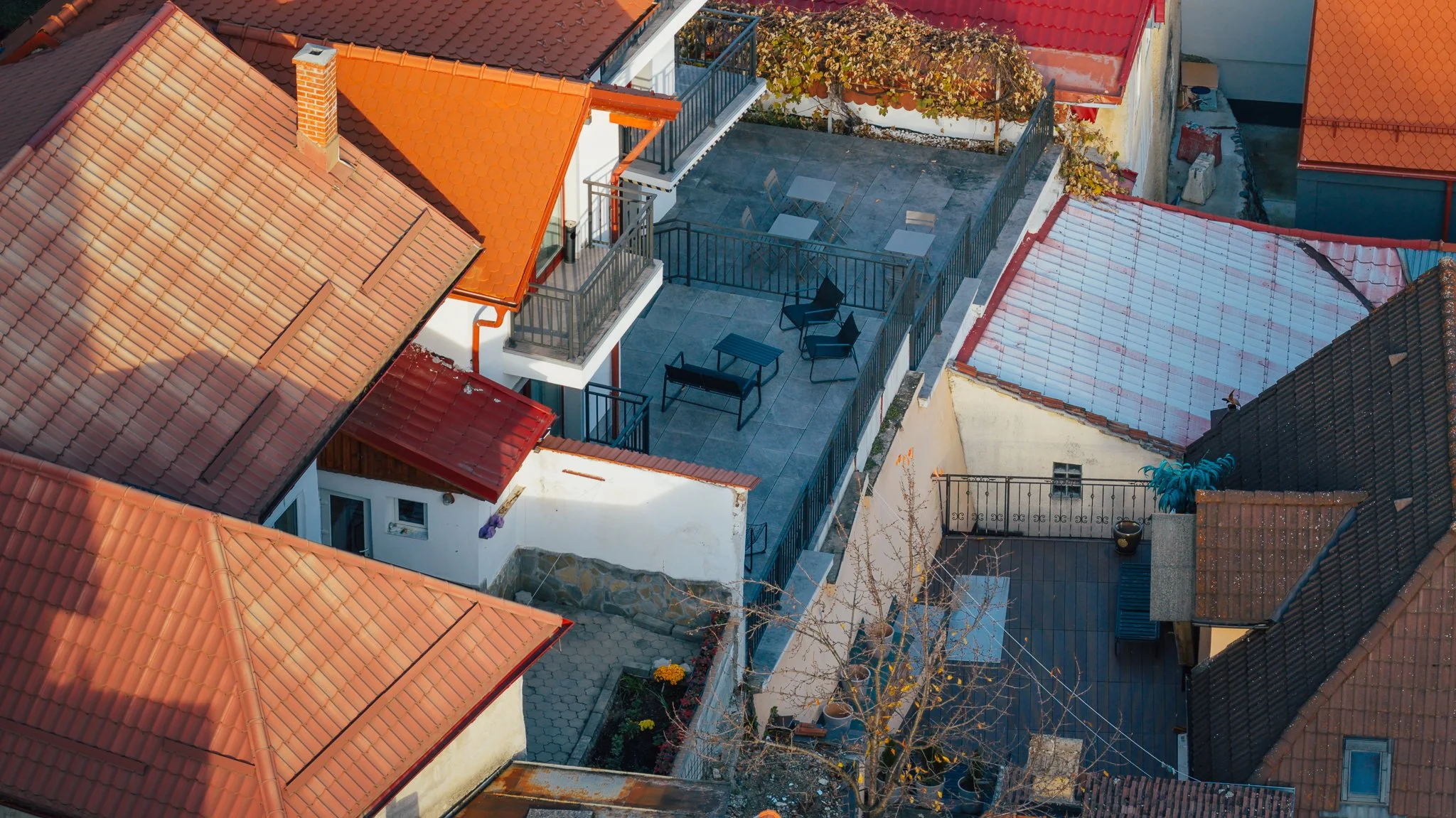 Aerial view of rooftops and balconies of multiple houses, with tiled roofs in red, orange, and brown, some with shingles, and a central terrace with outdoor furniture, enclosed by metal railings.