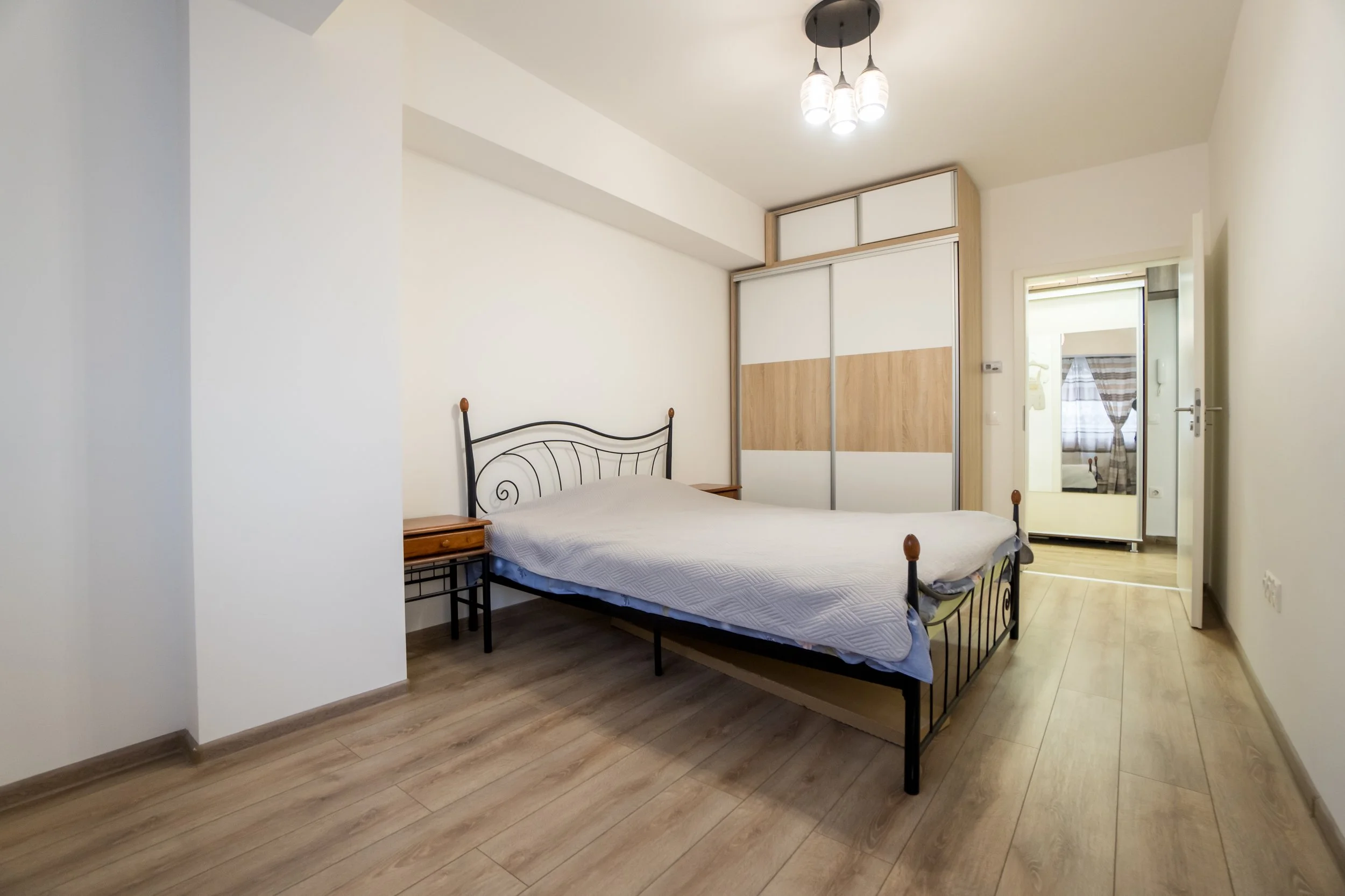 Bedroom with a metal bed frame, mattress, wooden nightstands, a sliding door wardrobe with wood and white panels, ceiling light fixture, wooden flooring, and a door leading to another room with curtains.