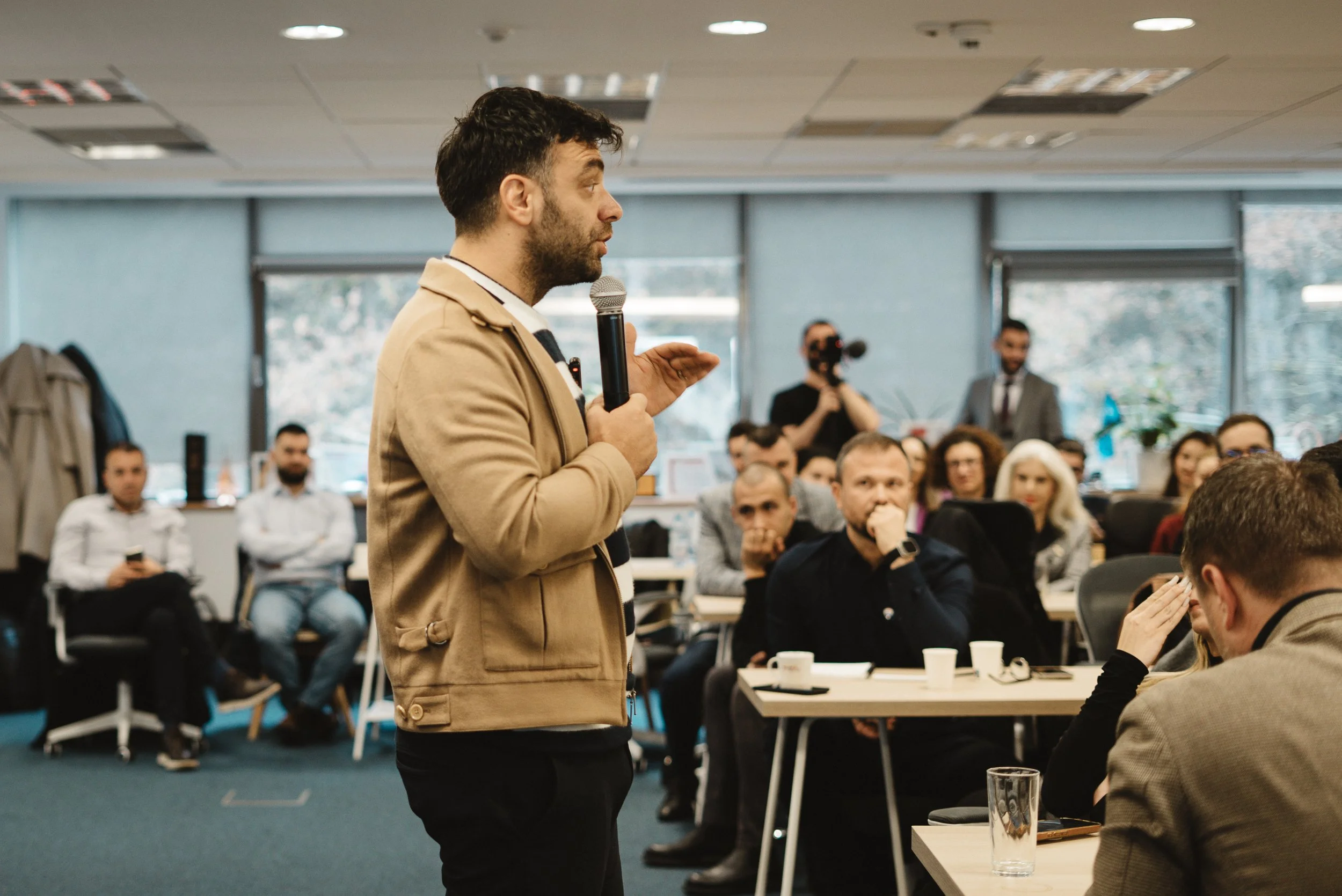 A man with dark hair and beard, wearing a beige jacket, speaking into a microphone in a conference room filled with seated attendees, some capturing the event on devices, with a photographer taking pictures in the background.