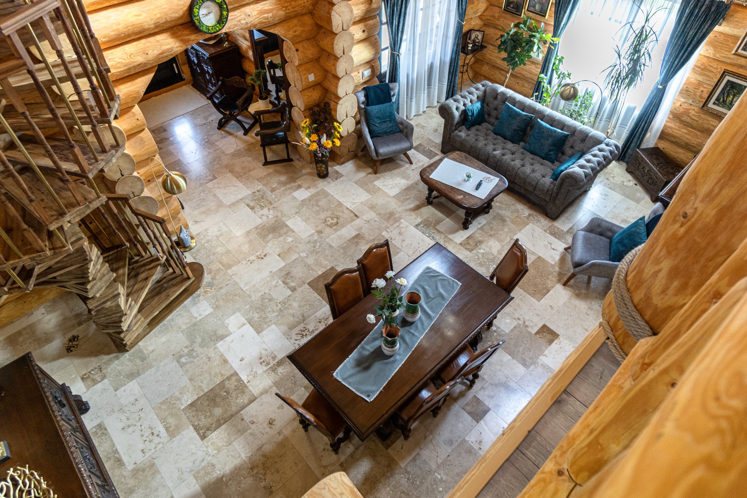 Living room and dining area inside a log cabin with wooden walls, a staircase, a dining table with chairs, a gray sofa, armchairs, plants, and large windows with curtains.