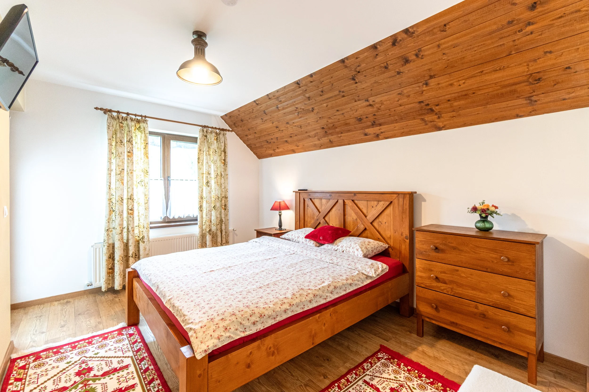 A cozy bedroom with a wooden bed, floral bedding, and a headboard made of wood. The room has a sloped wooden ceiling, a window with floral curtains, a small nightstand with a red lampshade, a wooden dresser with a flower vase, and a patterned red and
